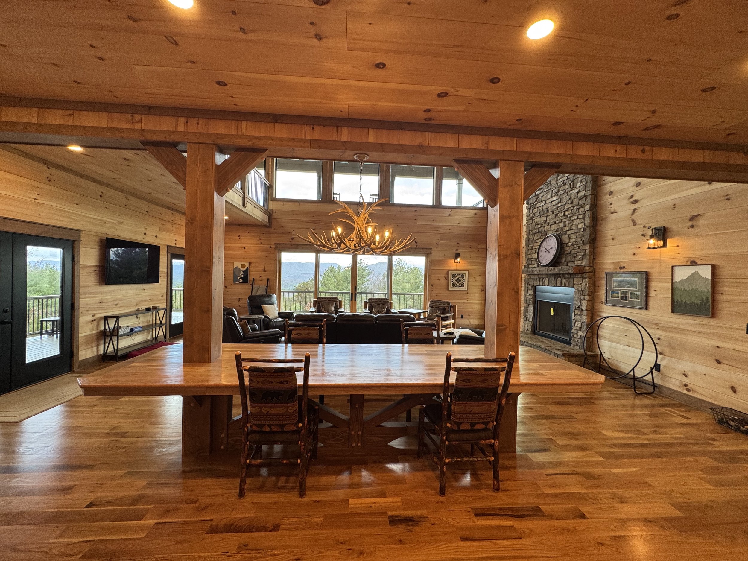 Spacious living and dining area in a cabin-style home with wooden walls and flooring, a large wooden dining table, leather chairs, a stone fireplace, and large windows offering mountain views.
