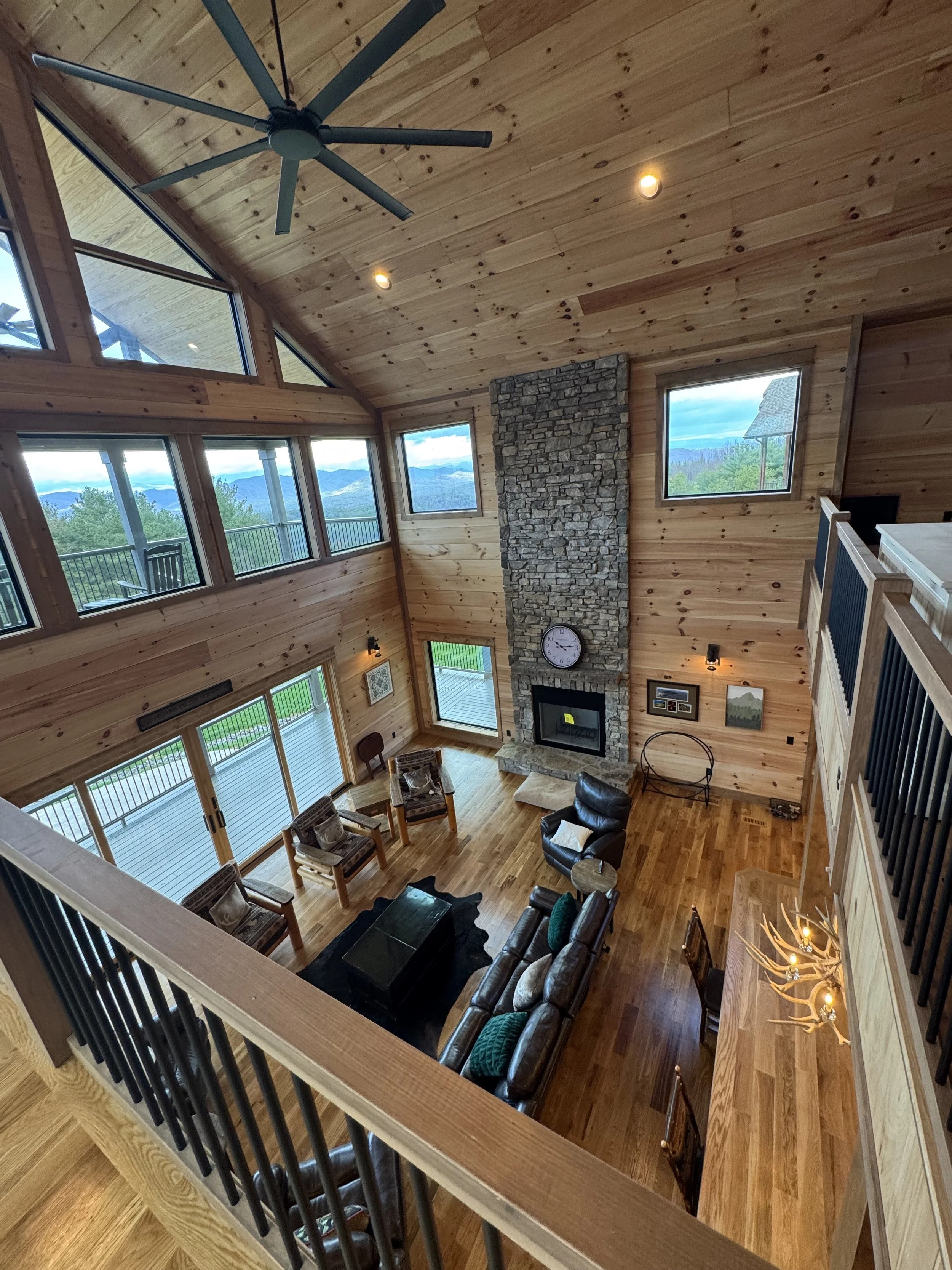 View of a spacious living room with vaulted wooden ceiling, large windows showing a mountain landscape, and a stone fireplace with a clock. The room features leather and wooden furniture, with a high balcony railing visible at the bottom.