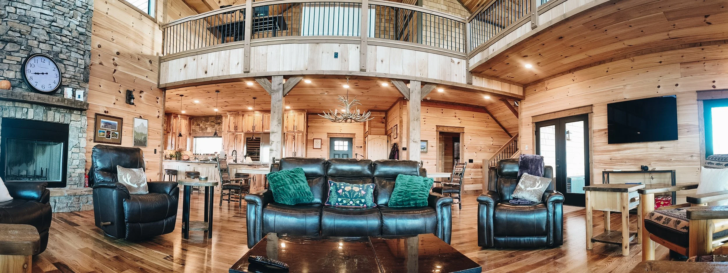A spacious, rustic living room with wood-paneled walls and ceiling, leather sofas with decorative pillows, a stone fireplace with a clock above, a mounted TV, and an open kitchen area in the background.