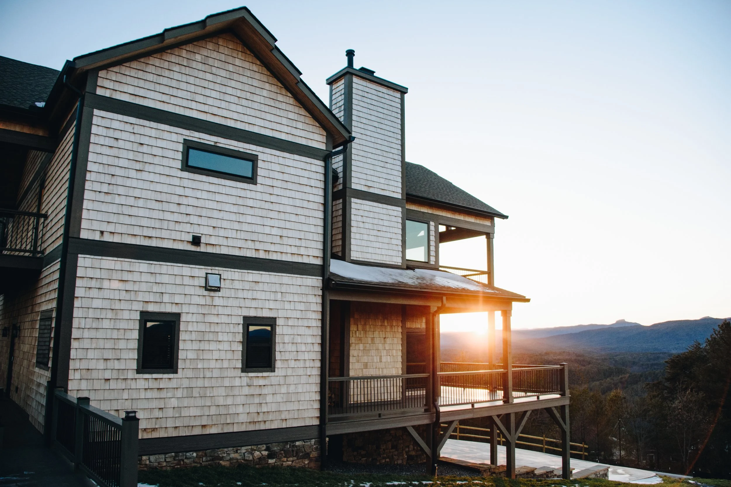 A multi-story house with wooden shingles and black trim, balcony, and large windows, with the sun setting behind mountains in the background.