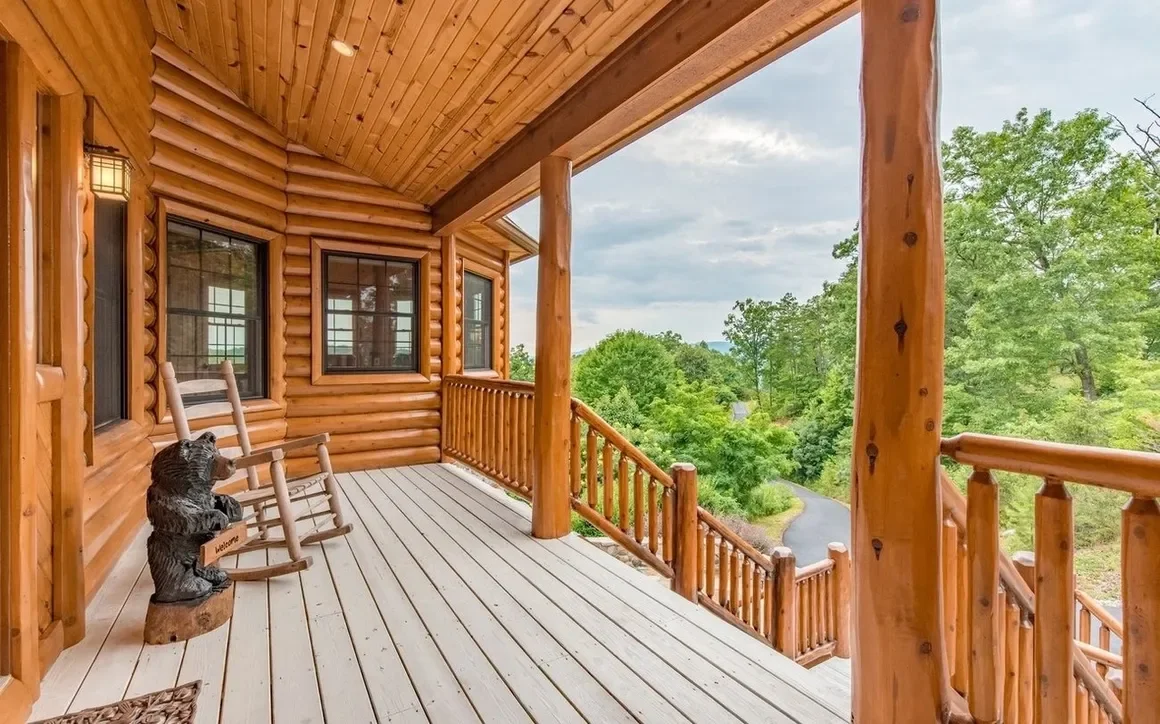 Wooden porch of a log cabin with rocking chairs and a bear sculpture, overlooking a lush green landscape with trees and a winding road.