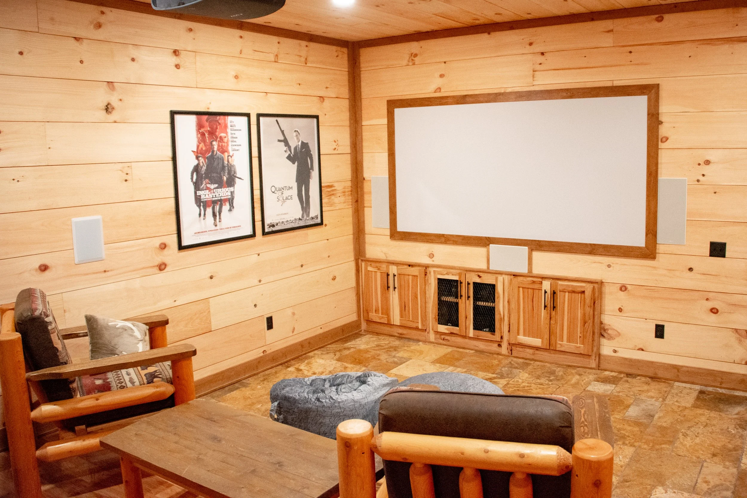 A cozy home theater room with wood-paneled walls, a large white projection screen, two framed movie posters on the wall, a wooden cabinet below the screen, and comfortable seating including an armchair and a sofa with pillows.