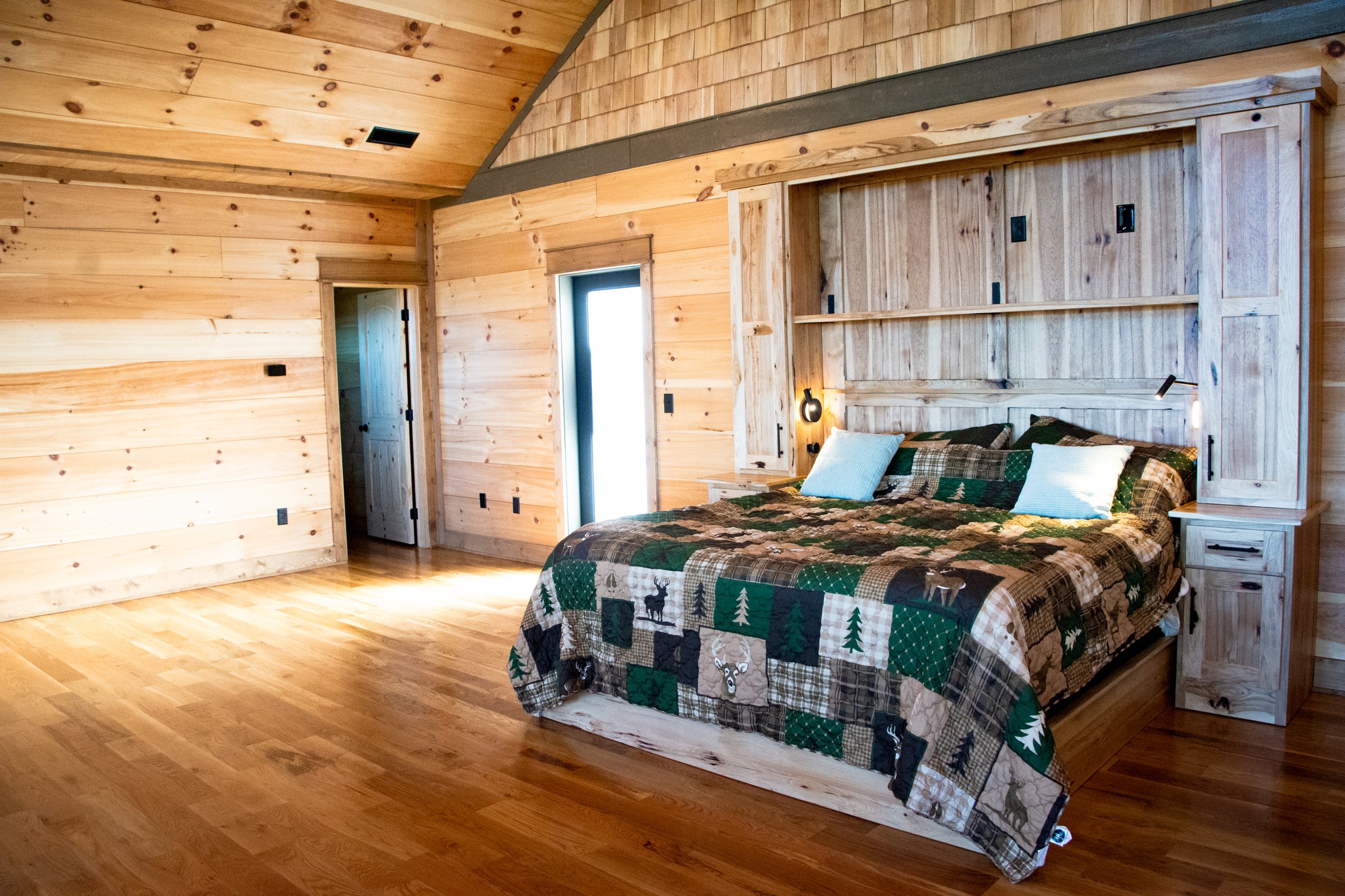 A cozy bedroom with wood-paneled walls, a large bed with a forest-themed quilt, and a small built-in bookshelf headboard. The room has natural light coming through a window and a door, with warm wooden flooring.