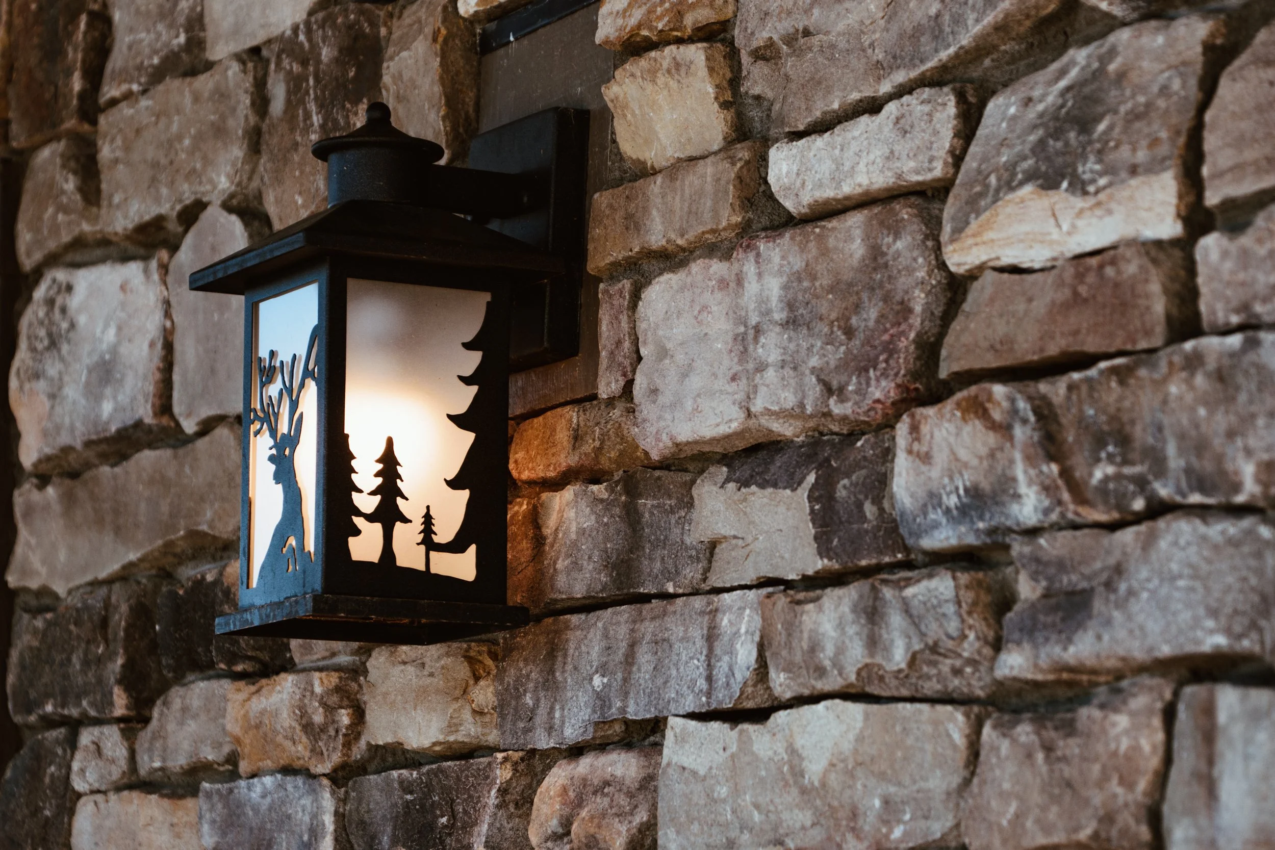 Black outdoor wall lantern with a mountain scene silhouette design, mounted on a stone brick wall.