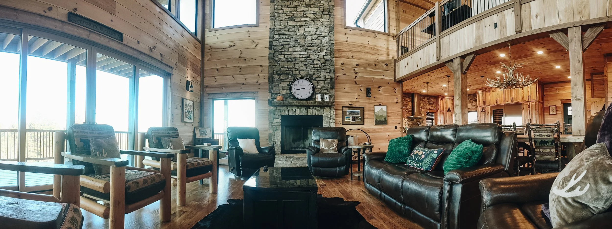 Living room with large windows, stone fireplace, wooden walls, leather sofa, armchairs, coffee table, and rustic decor.