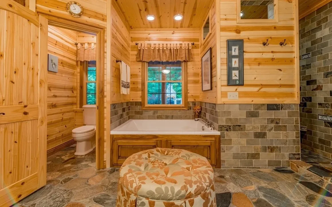 A rustic bathroom with wooden walls, a bathtub beneath a window, a toilet in a separate room, and a stone floor.