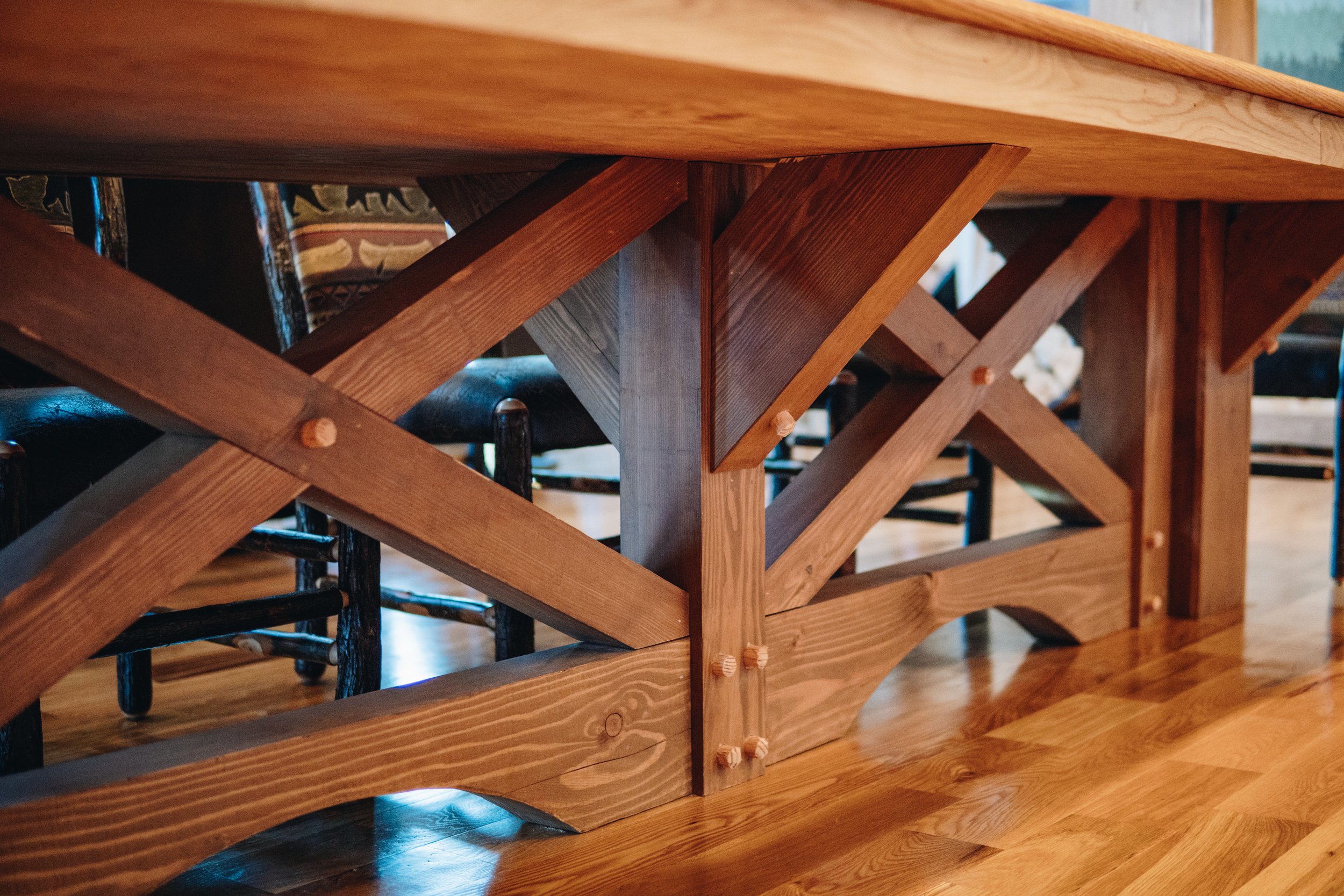 Close-up view of a wooden table’s underside with intersecting wooden supports and bolts, showing a sturdy construction with a natural wood finish, resting on a wood floor.