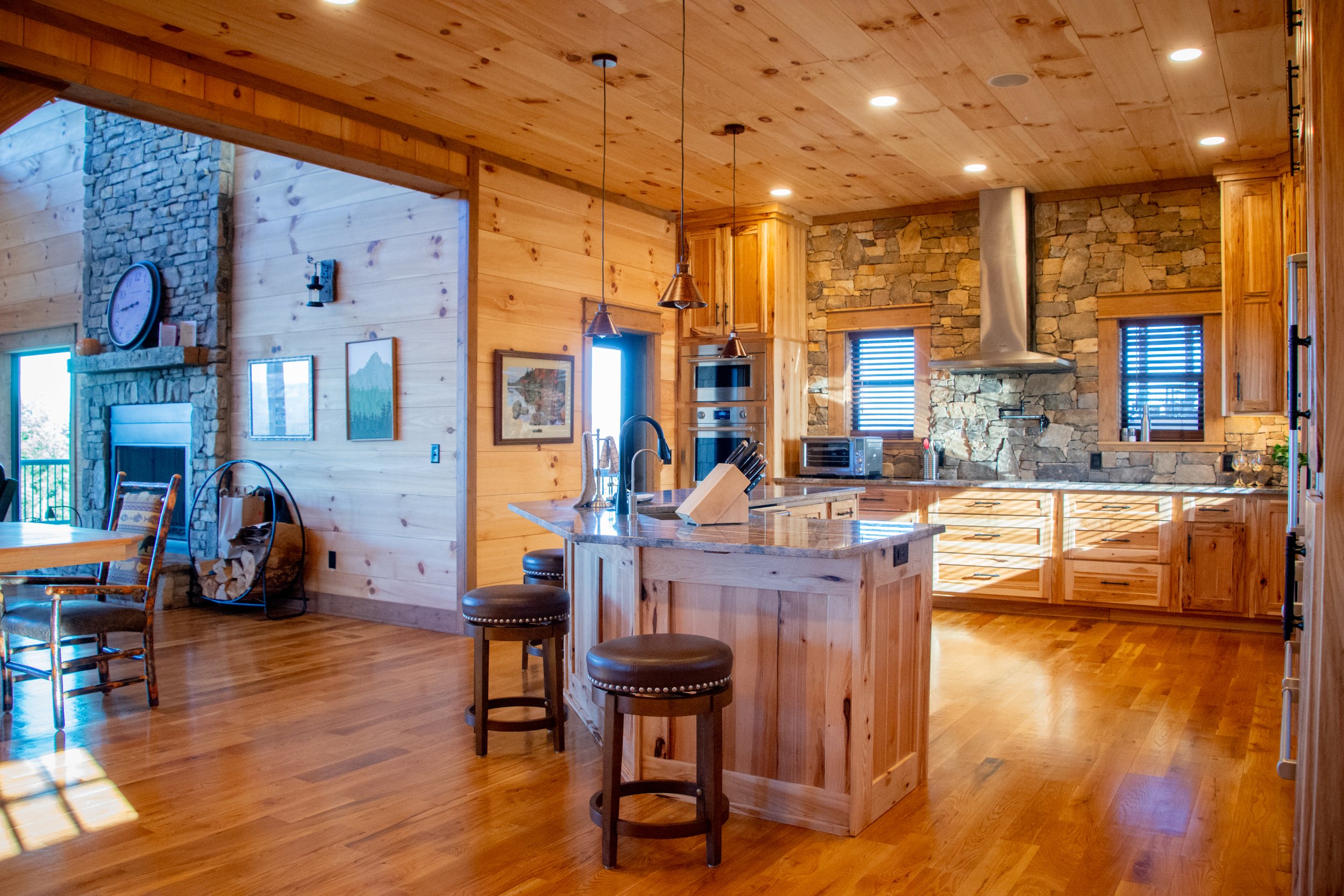 Rustic kitchen with wooden cabinets, stone wall, island with sink and stools, stainless steel range hood, and natural light coming through shutters.