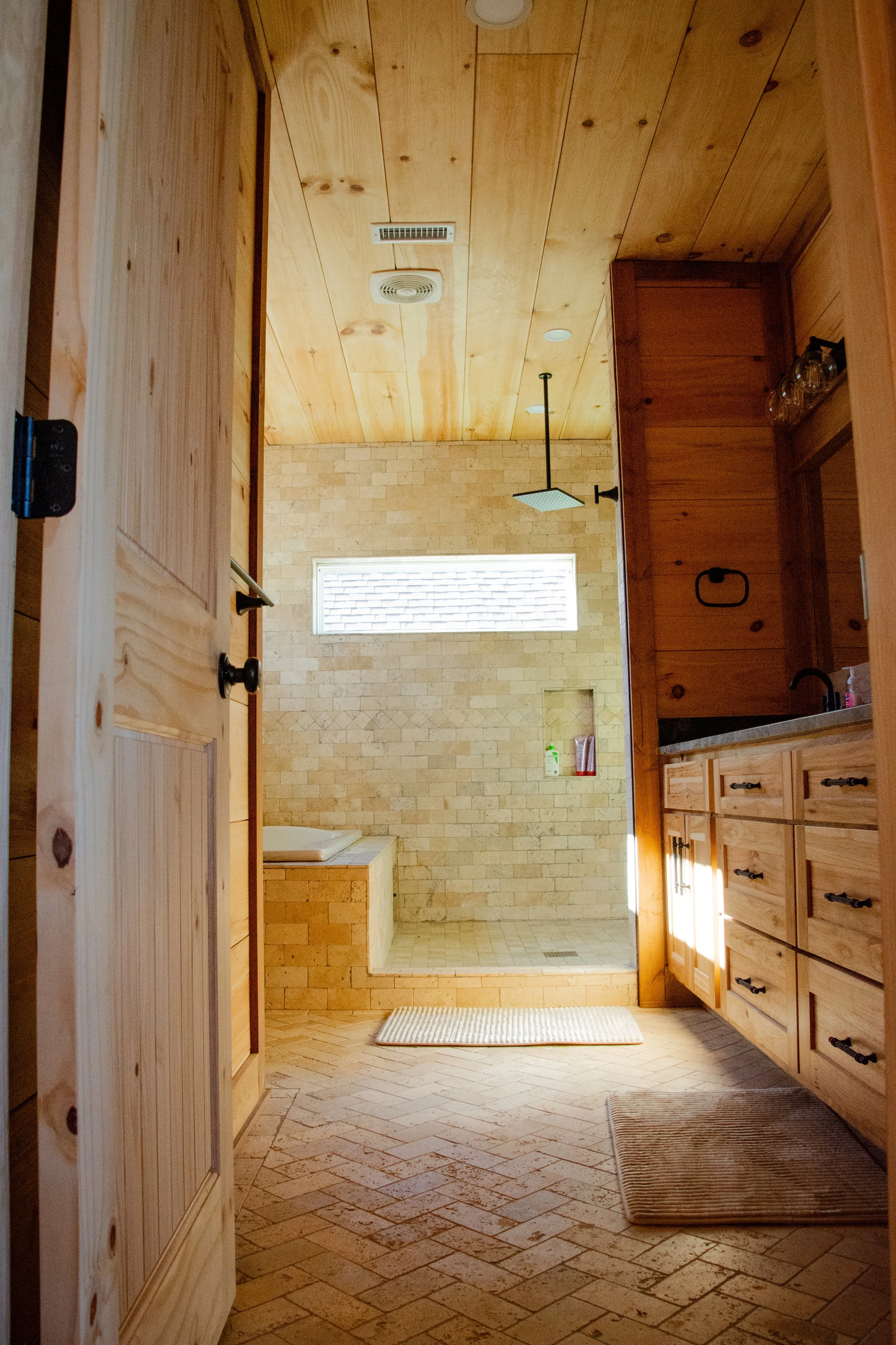 Interior of a bathroom with wooden cabinets, brick shower walls, a built-in bathtub, and small rectangular window letting in natural light.