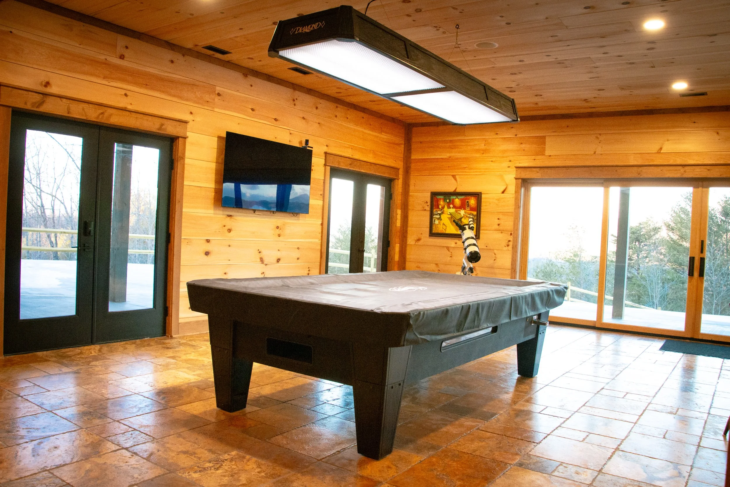 A game room with wood-paneled walls and ceiling, large sliding glass doors leading outside, a pool table covered with a gray cover, a wall-mounted TV, a telescope, and a colorful painting.