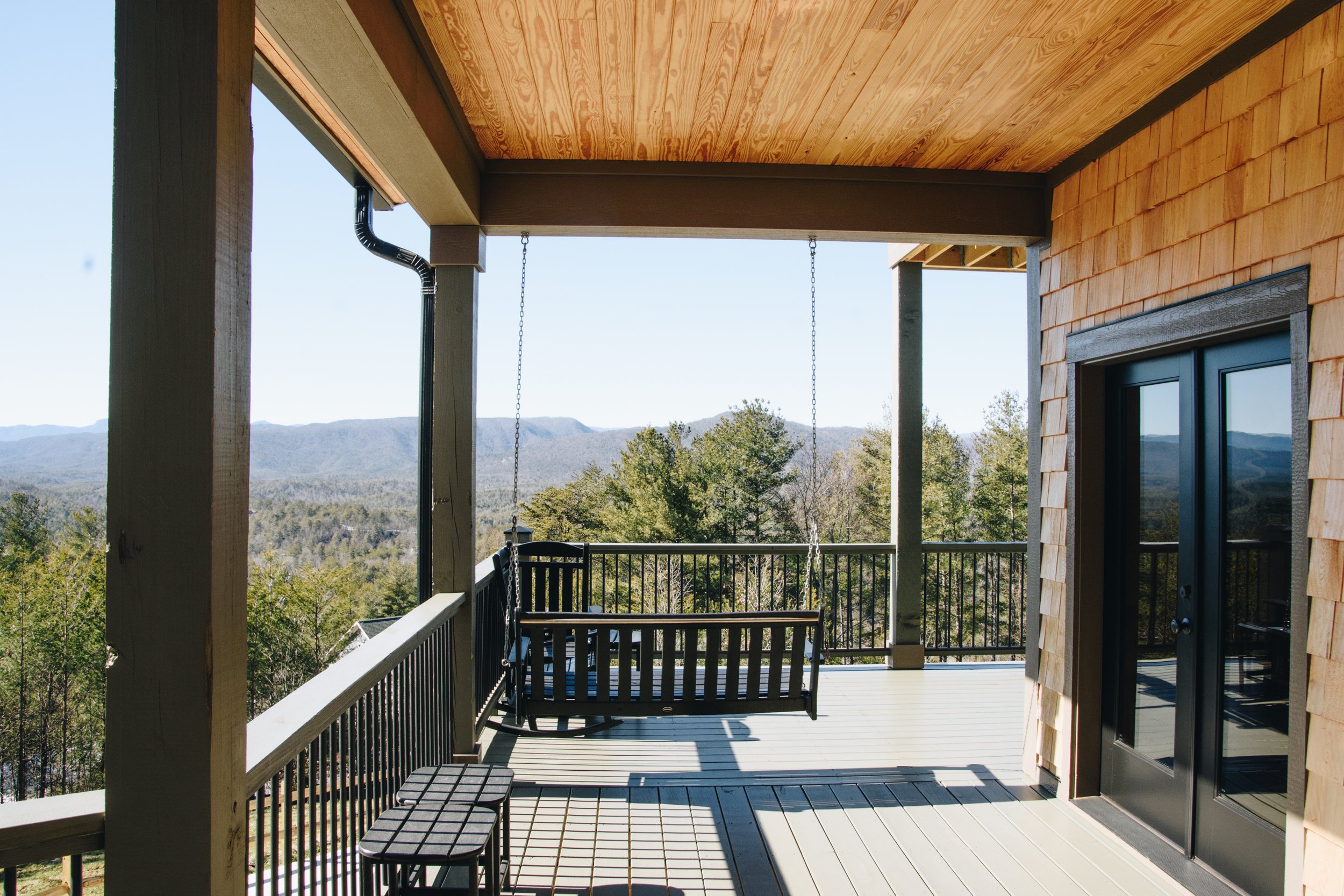 A wooden balcony with black railings, a swing seat, a small table with stools, and a mountain view in the background.