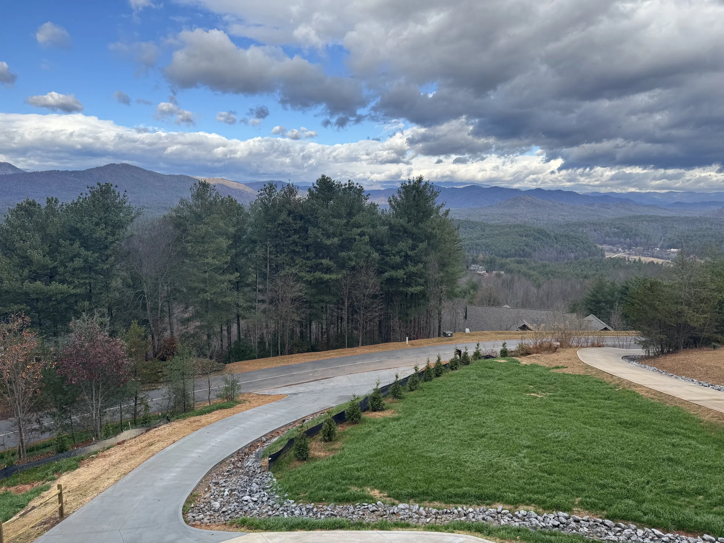A landscaped yard with green grass, a curved concrete path, and small shrubs, with wooded hills and mountains in the background under a partly cloudy sky.