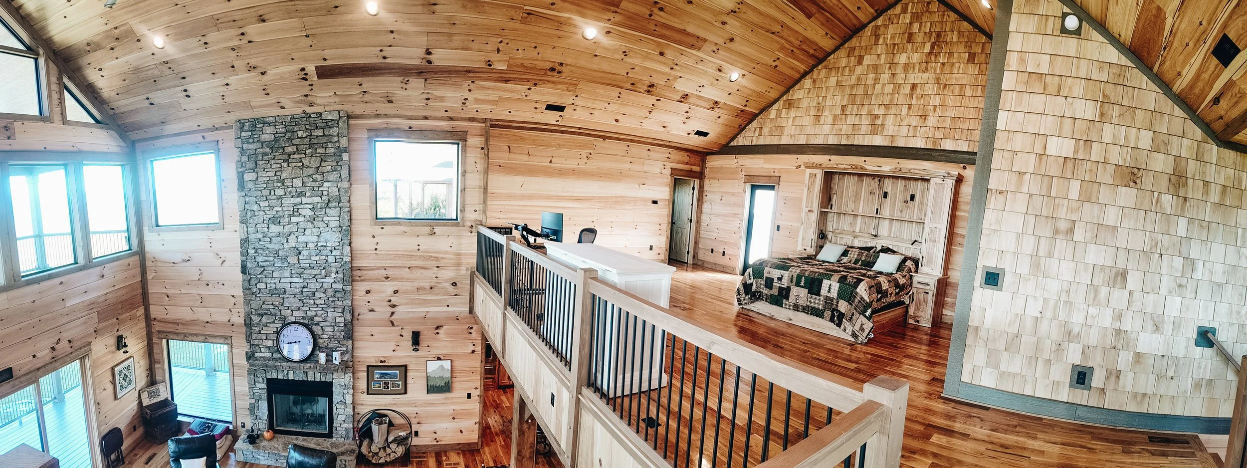 Interior view of a wooden home with a high vaulted ceiling, large windows, a stone fireplace, a sitting area with a couch, and a loft area with a desk and chair.