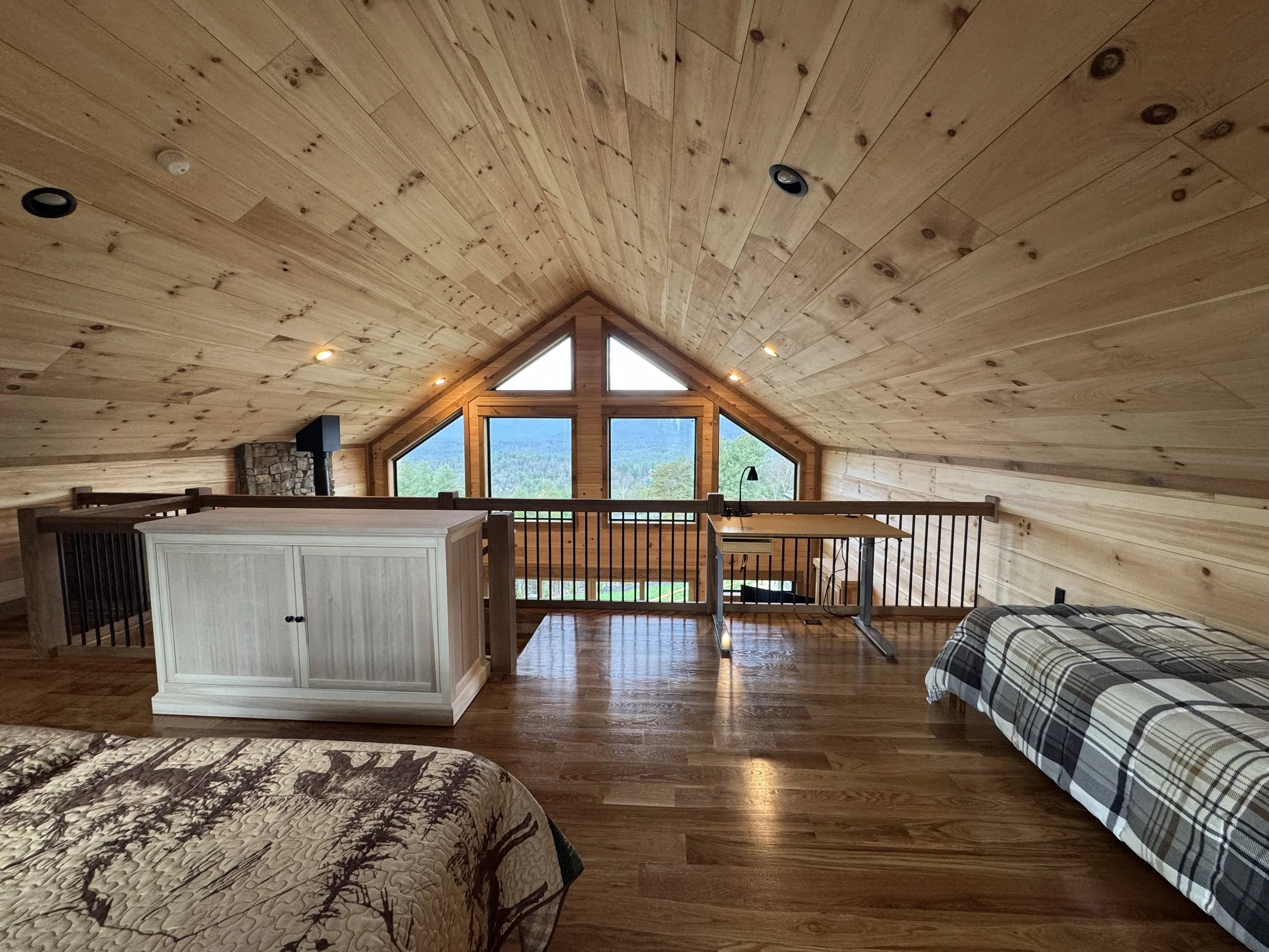 Loft bedroom with sloped wooden ceiling, large triangular window showing mountain and forest views, and two beds with plaid and deer-themed bedding.