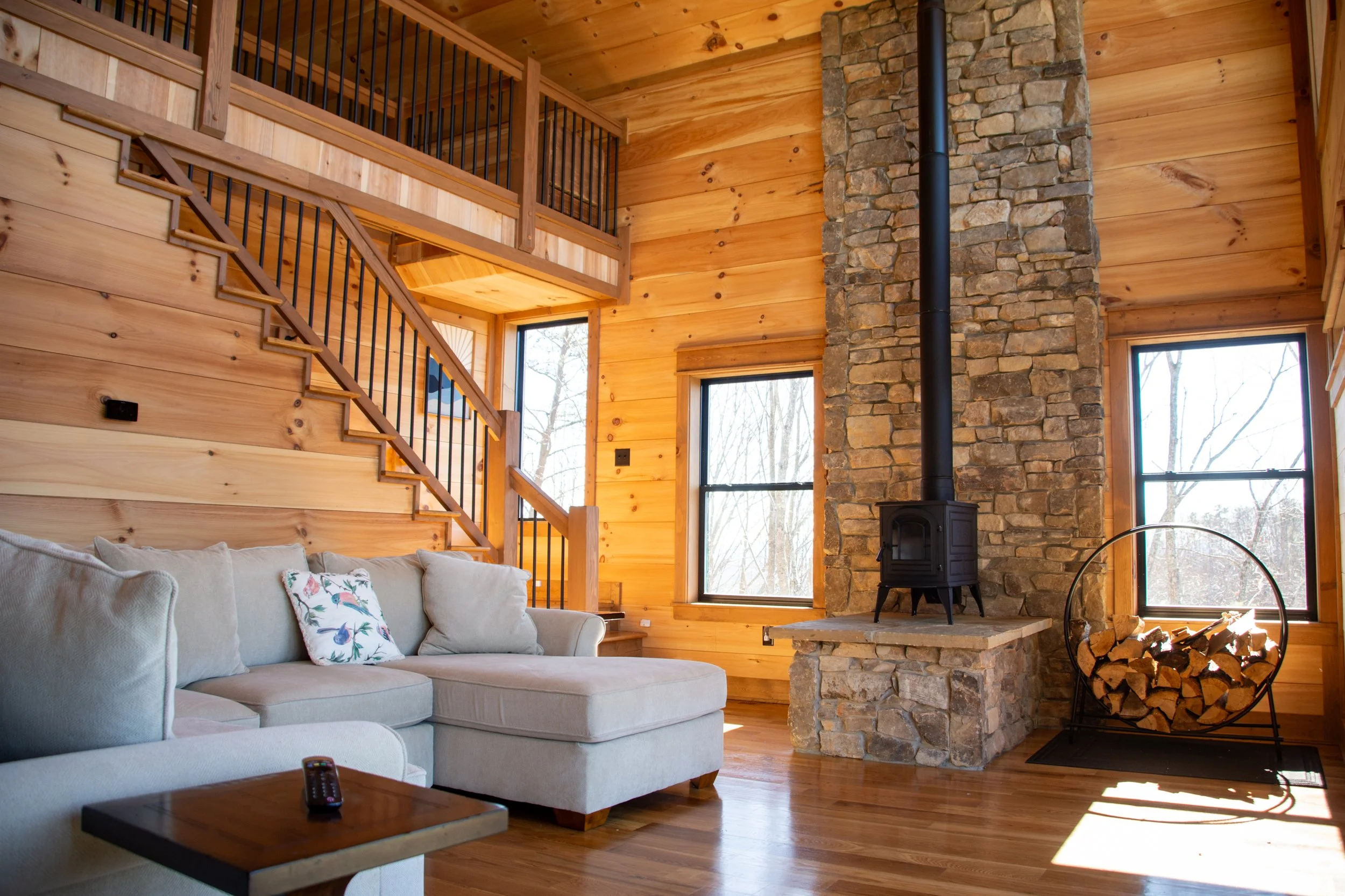 Cozy living room with wooden walls, a beige sofa with floral pillow, a wood stove on a stone chimney, large windows, and a wood log holder filled with firewood.