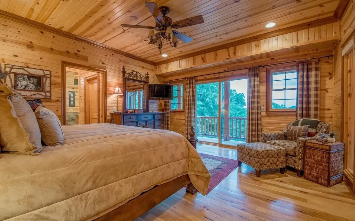 A cozy bedroom with wooden walls and ceiling, a large bed with beige bedding, a ceiling fan, a sliding glass door leading to a balcony, and a sitting area with an armchair and ottoman.