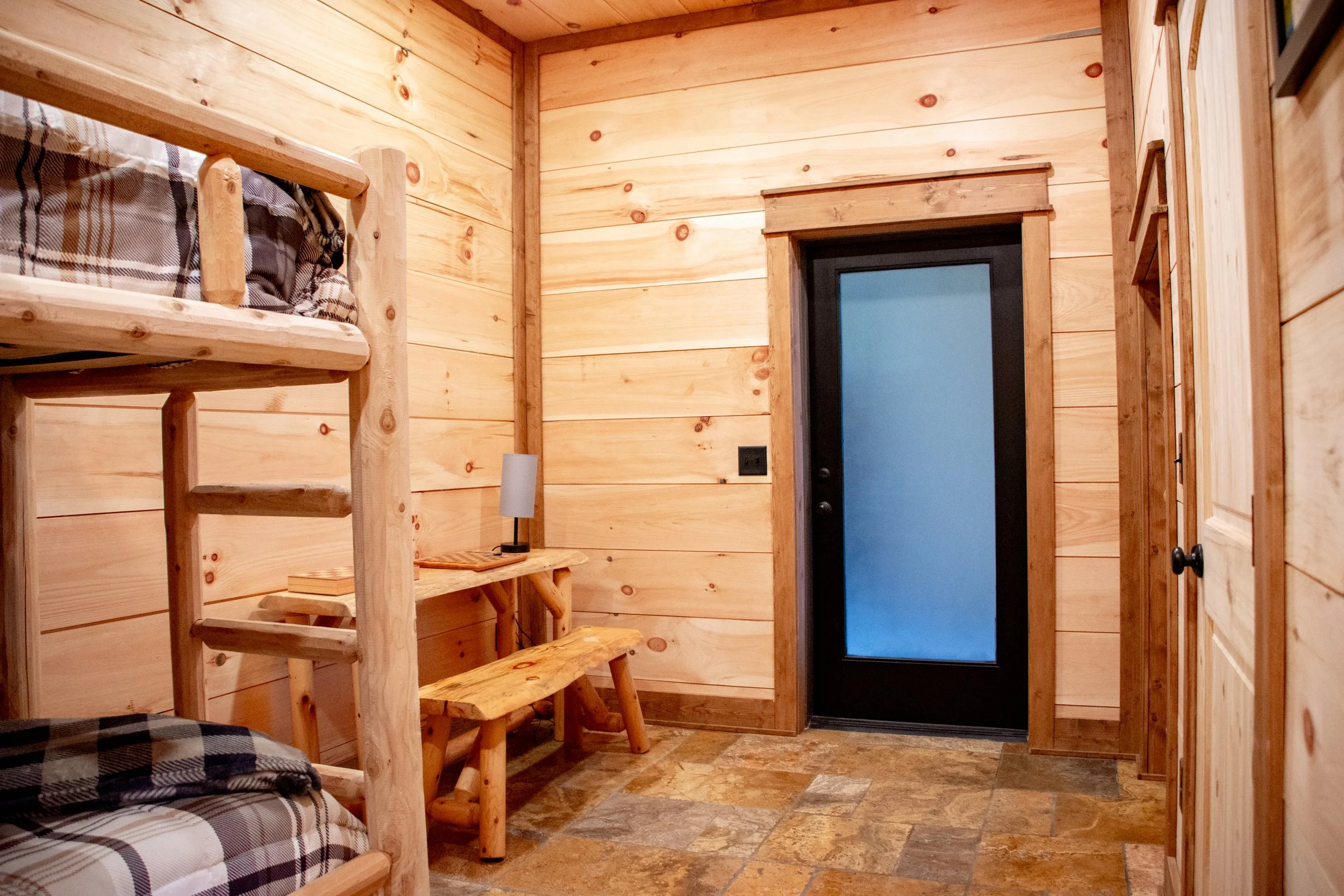 A small wooden room with a bunk bed, a wooden bench, a small wooden desk with a lamp on it, and a black door with frosted glass, all with wood paneling walls and a stone floor.