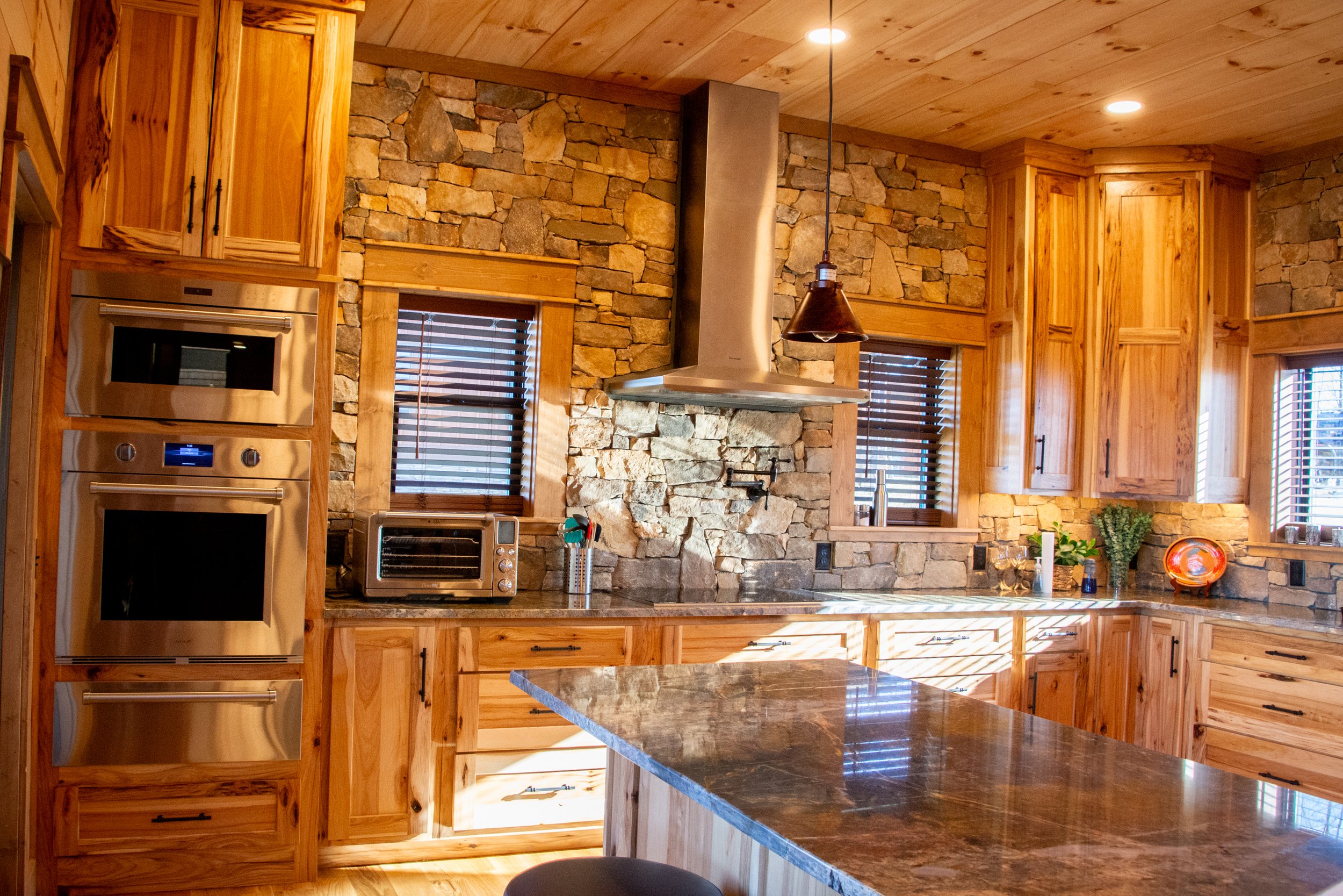 A kitchen with wooden cabinets, stone walls, stainless steel appliances, and a granite island.