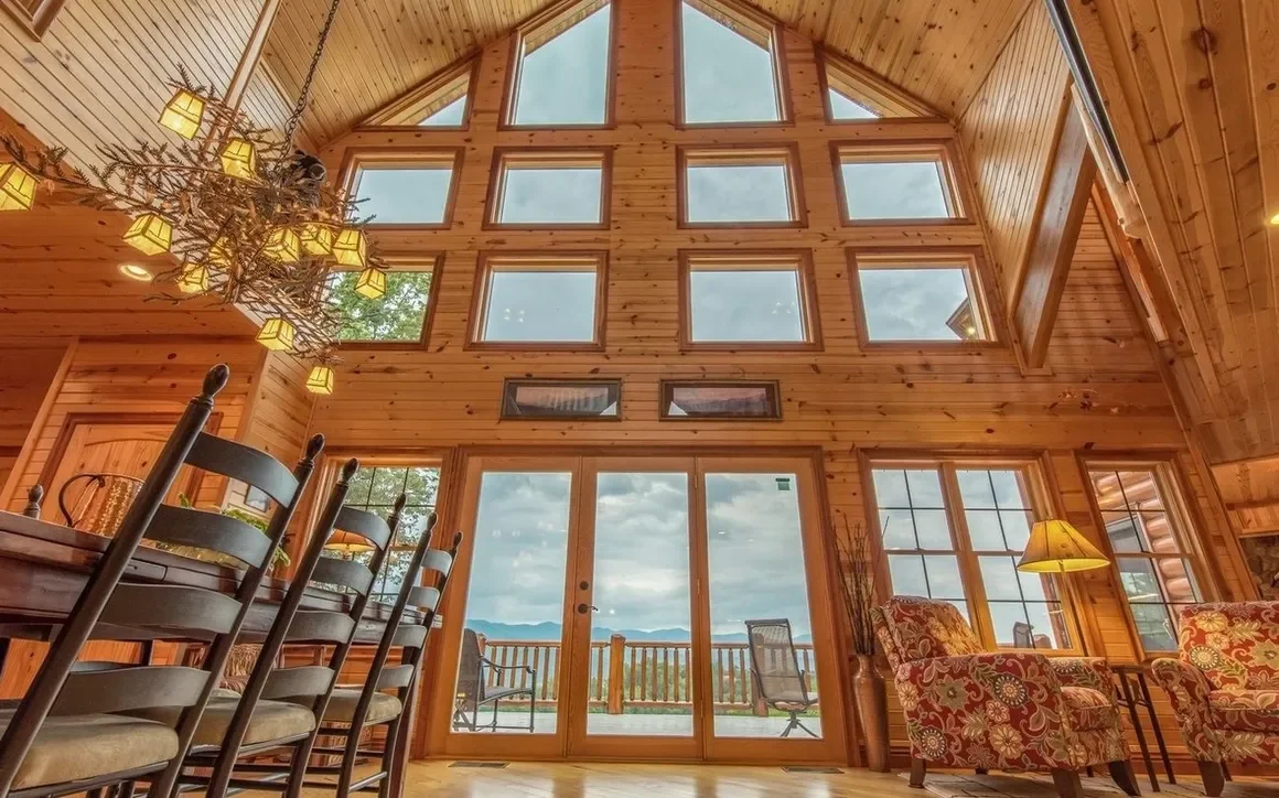 Interior of a wooden cabin with large windows, a chandelier, chairs, armchairs with floral patterns, and a sliding glass door leading to an outdoor deck with mountain views.