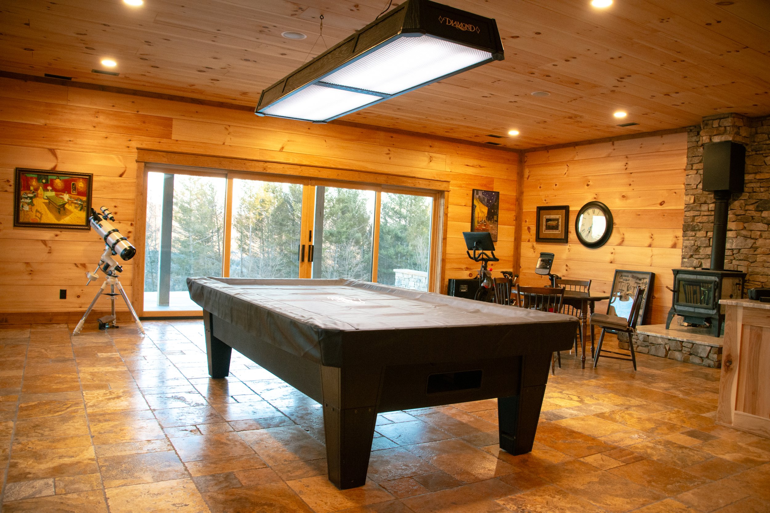 A room with wooden walls and ceiling, featuring a pool table, a telescope, several chairs, a fireplace, and sliding glass doors leading outside to a wooded area.