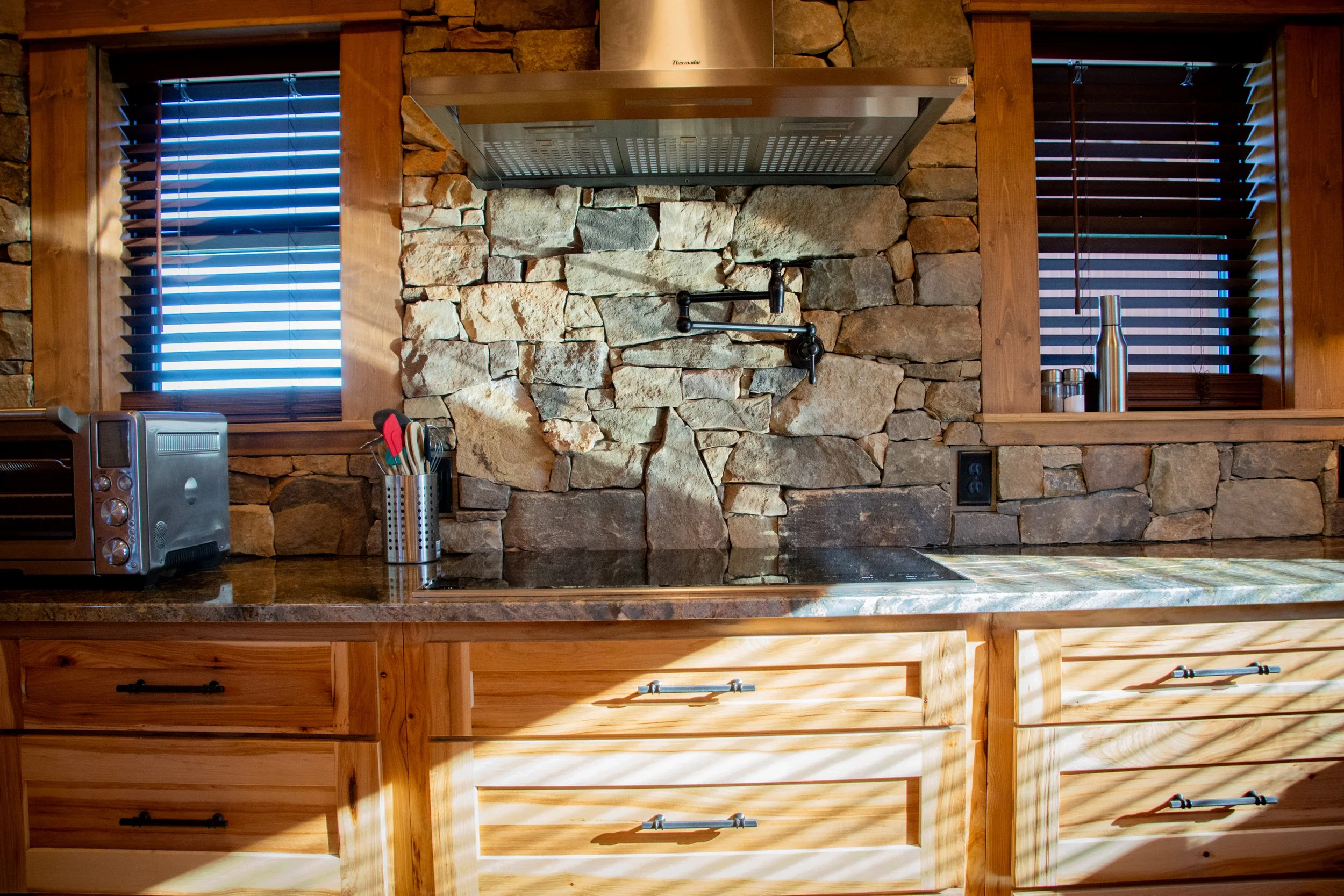 A rustic kitchen with wooden cabinets, a stone backsplash, and two windows with wooden blinds. On the countertop, there is a toaster, a container with utensils, and a stainless steel thermos. The kitchen has warm wood tones and a modern stainless ste