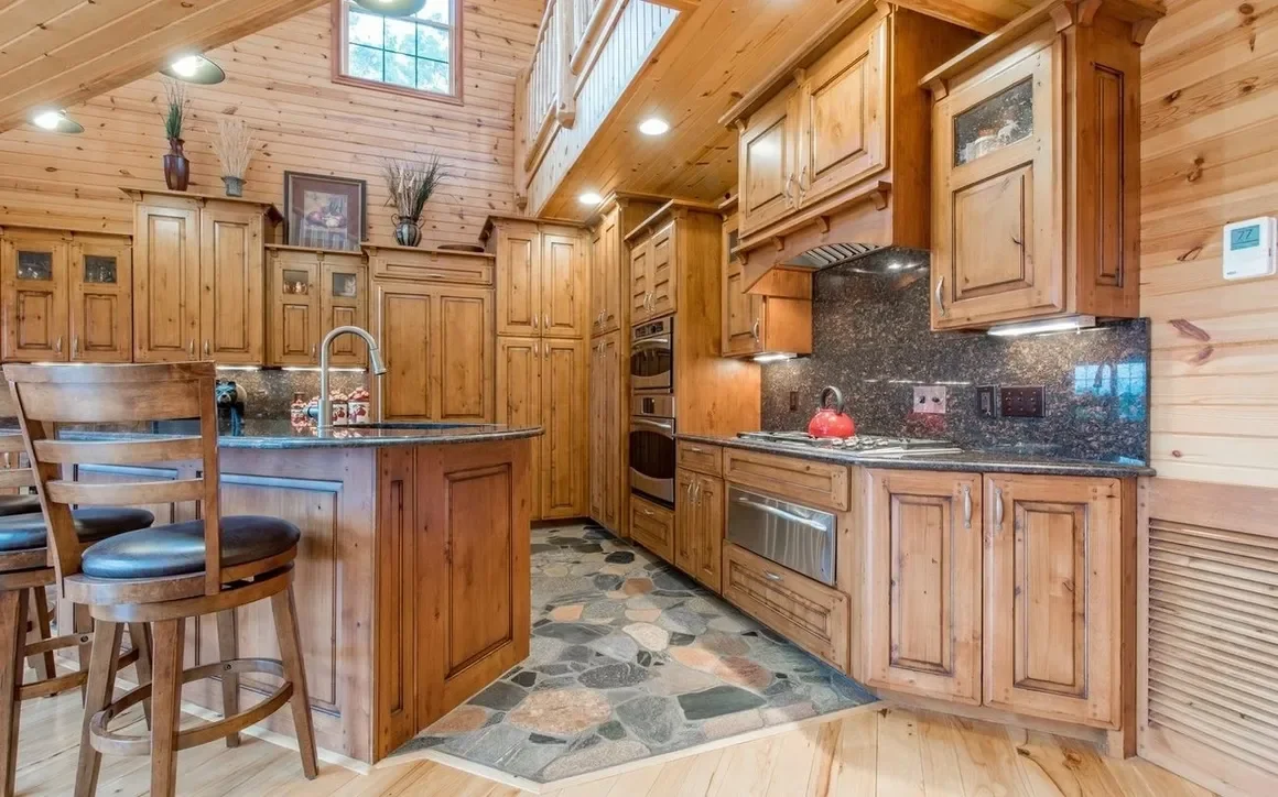 Wooden kitchen with granite countertops, cabinets, and island, featuring double ovens, a stove with a red kettle, wooden chairs, and a stone floor.