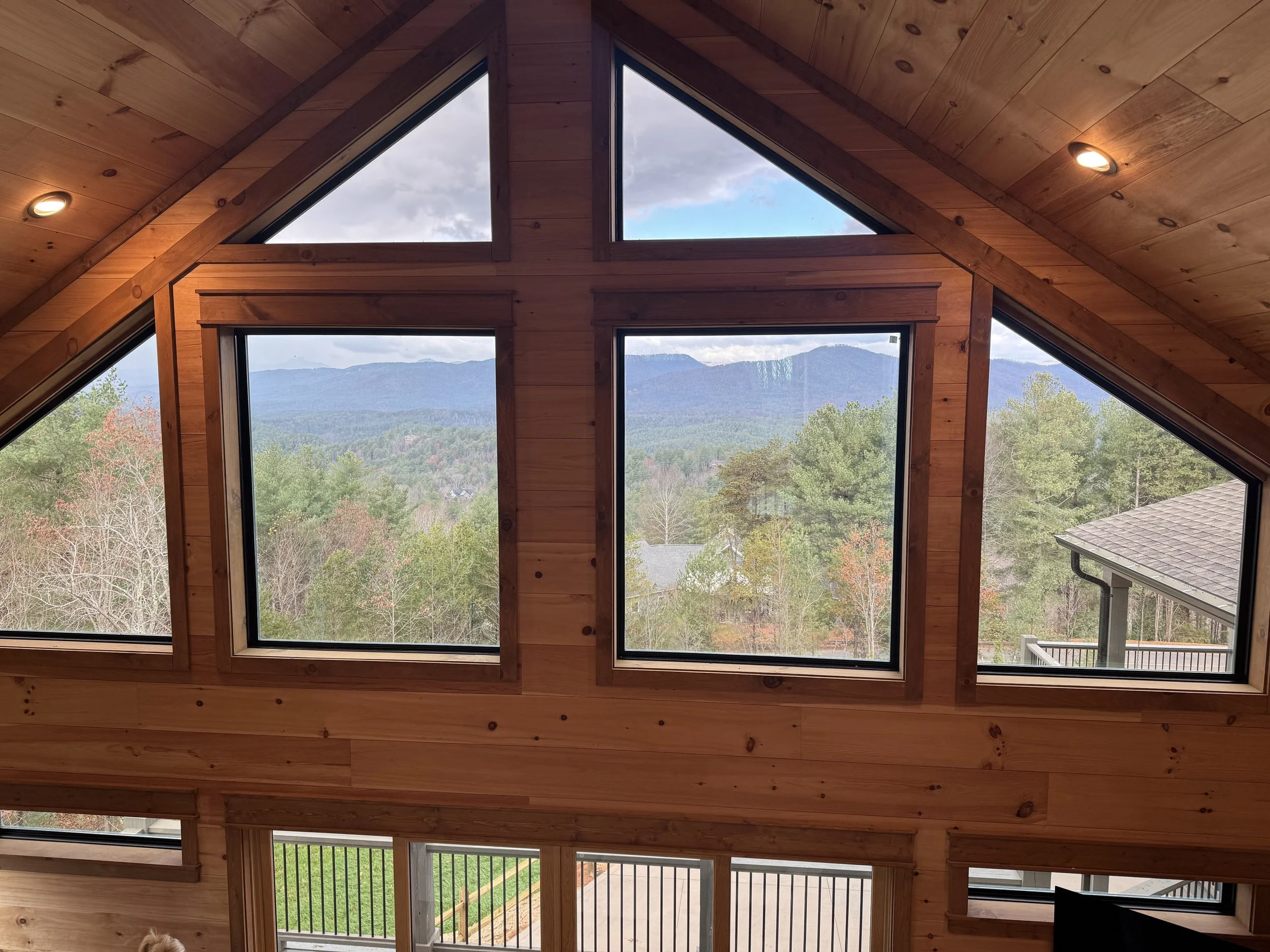 Interior of a wooden house with large triangular and rectangular windows, showing a scenic view of mountains and trees outside.
