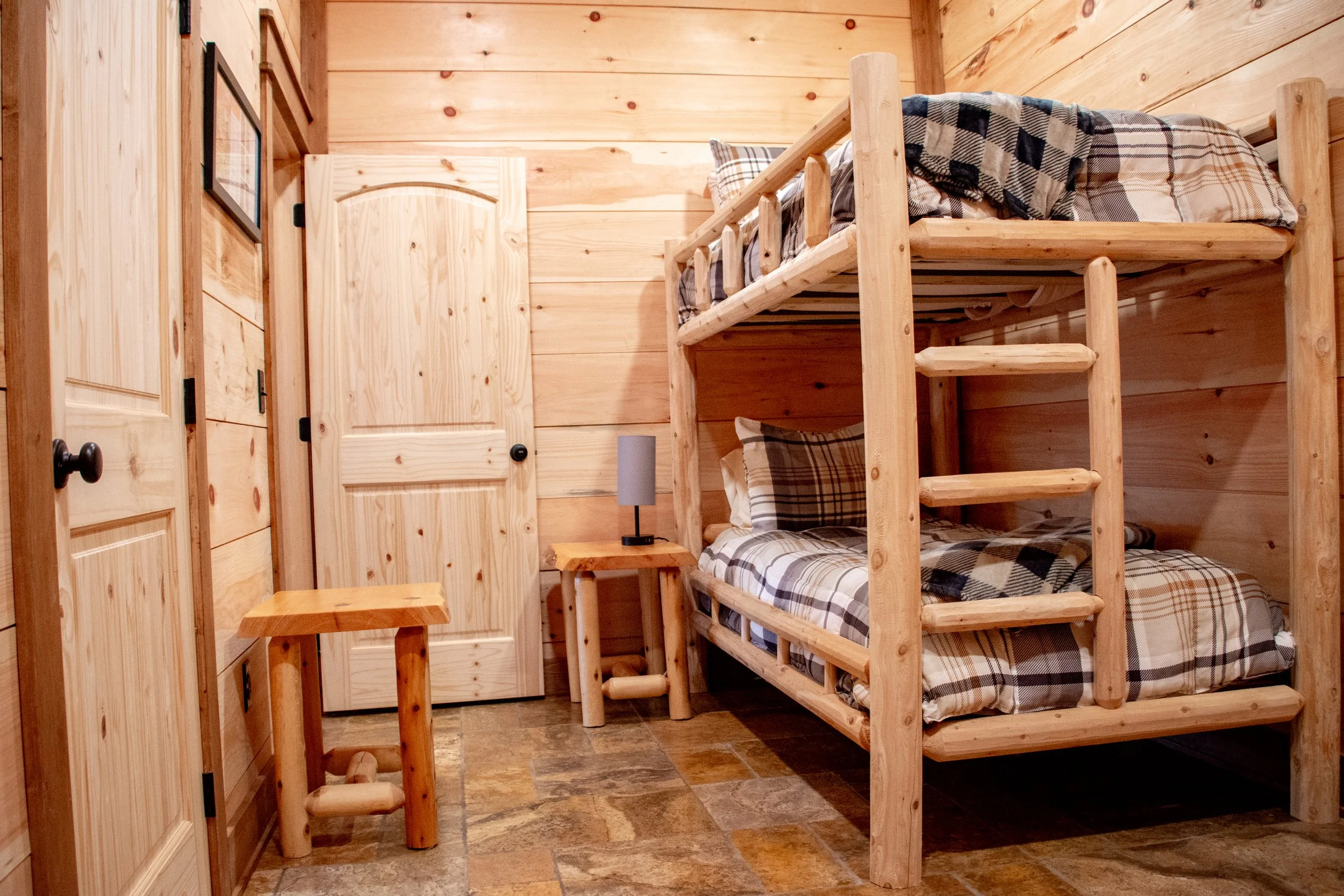A wooden bunk bed with plaid bedding in a rustic cabin room, with small tables and paintings on wooden walls.