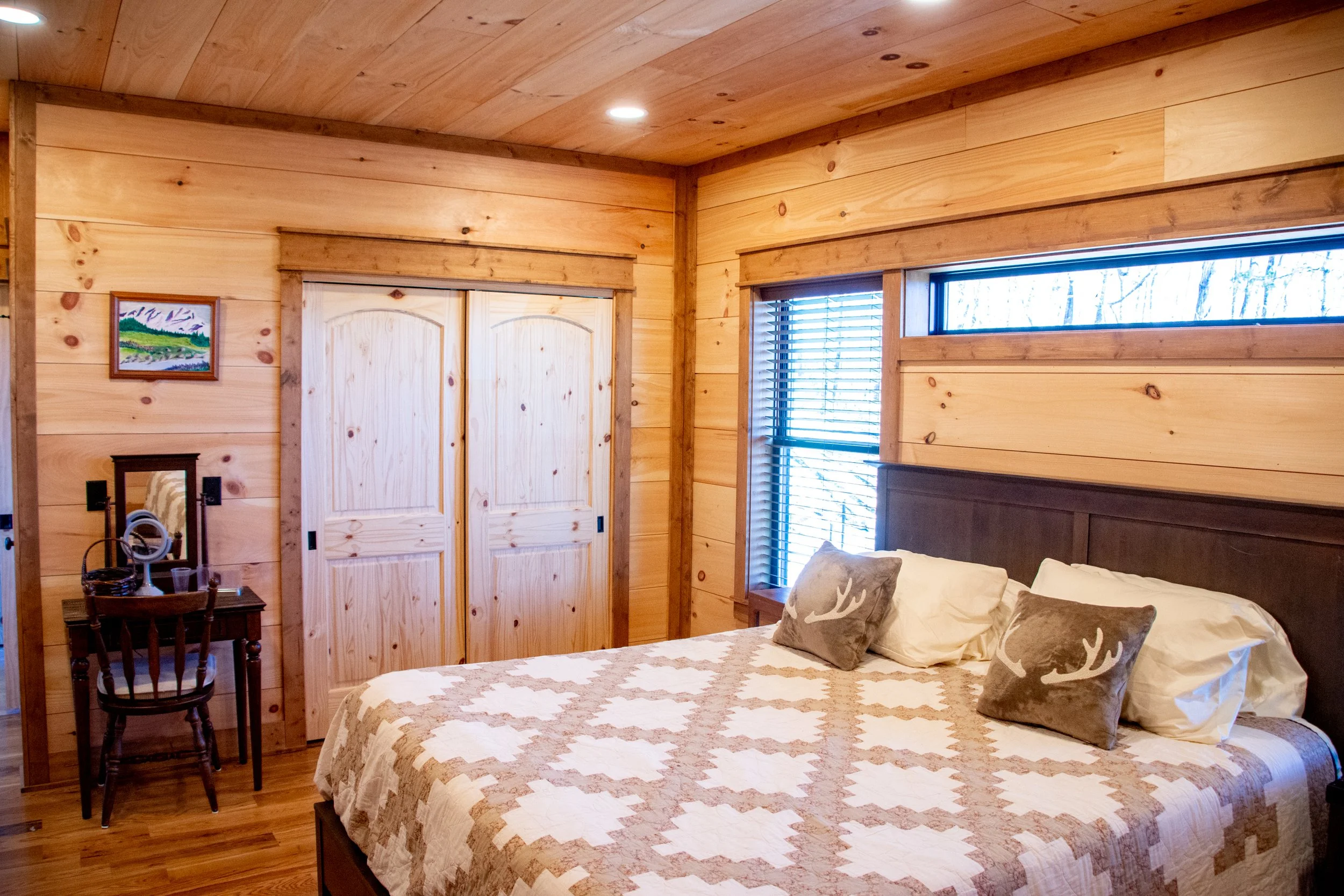 Cozy bedroom with wood-paneled walls, a bed with a quilted bedspread and decorative pillows, a small wooden desk with a mirror, a painting on the wall, a closet with sliding doors, and a window with blinds.