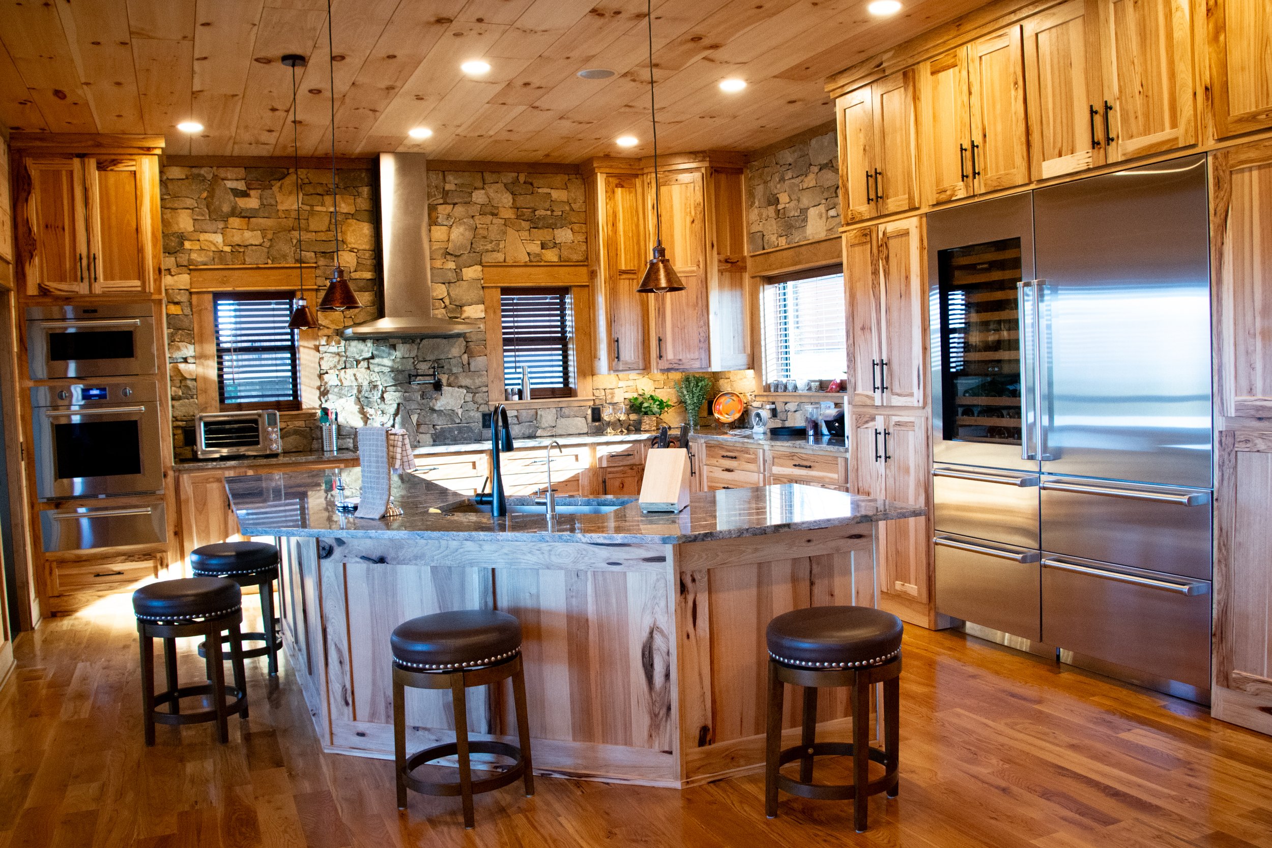 Rustic kitchen with wooden cabinetry, stone wall, stainless steel appliances, granite island, and hardwood floors.