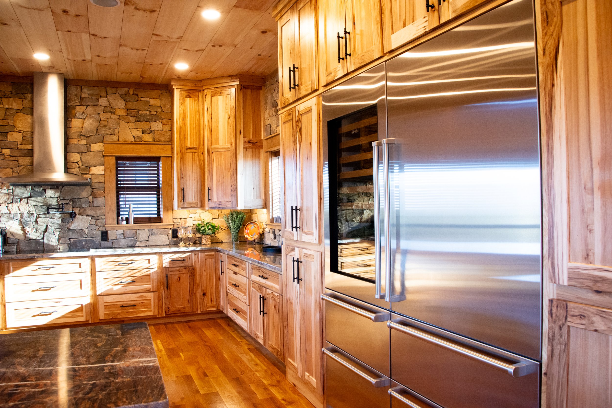 A rustic kitchen with wooden cabinets, stone walls, a stainless steel refrigerator, and a marble countertop. There are small windows with blinds and some decorative plants on the countertop.