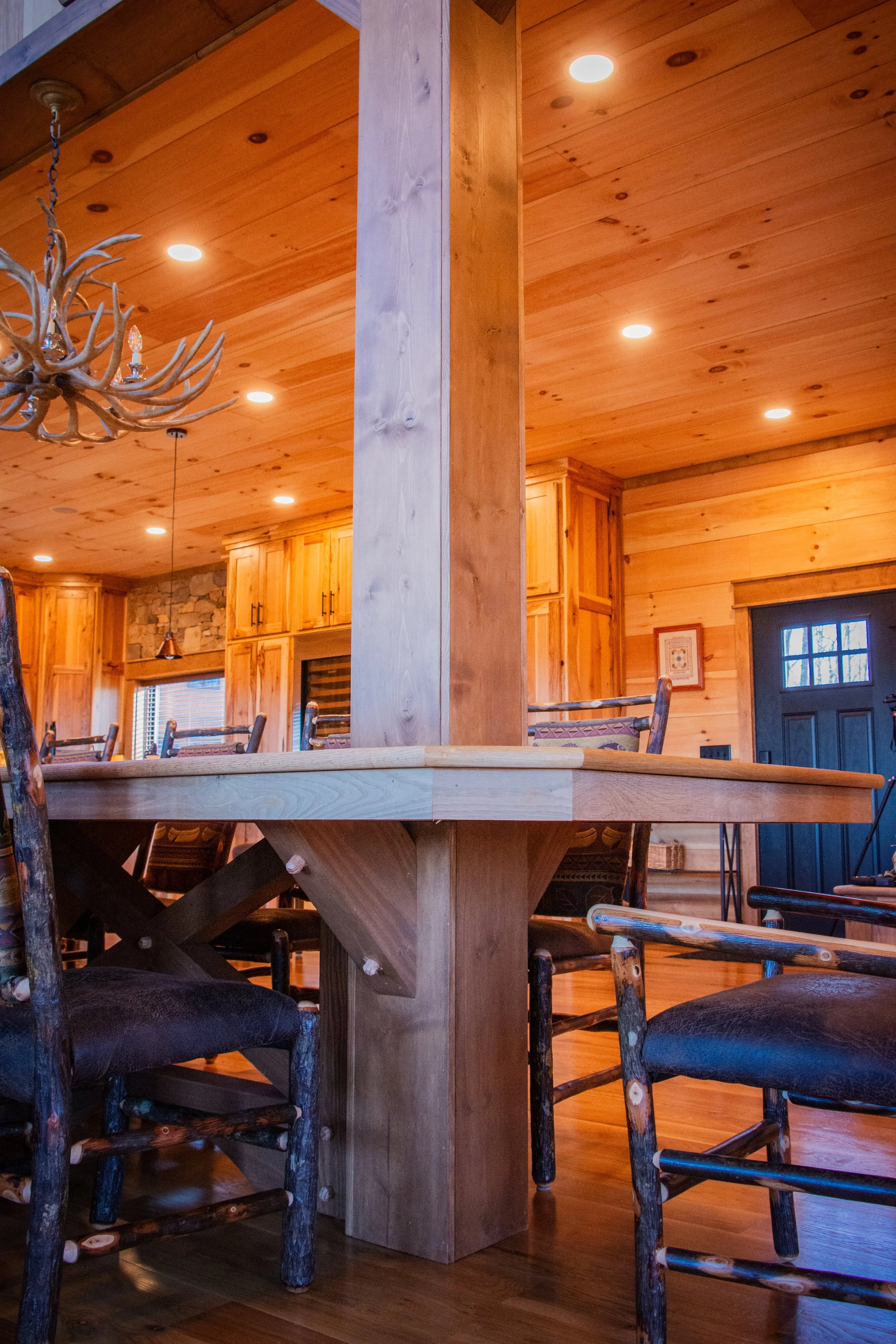 Interior of a rustic wood cabin with a large wooden dining table, surrounding chairs, pendant and chandelier lighting, and wood panel walls and ceiling.