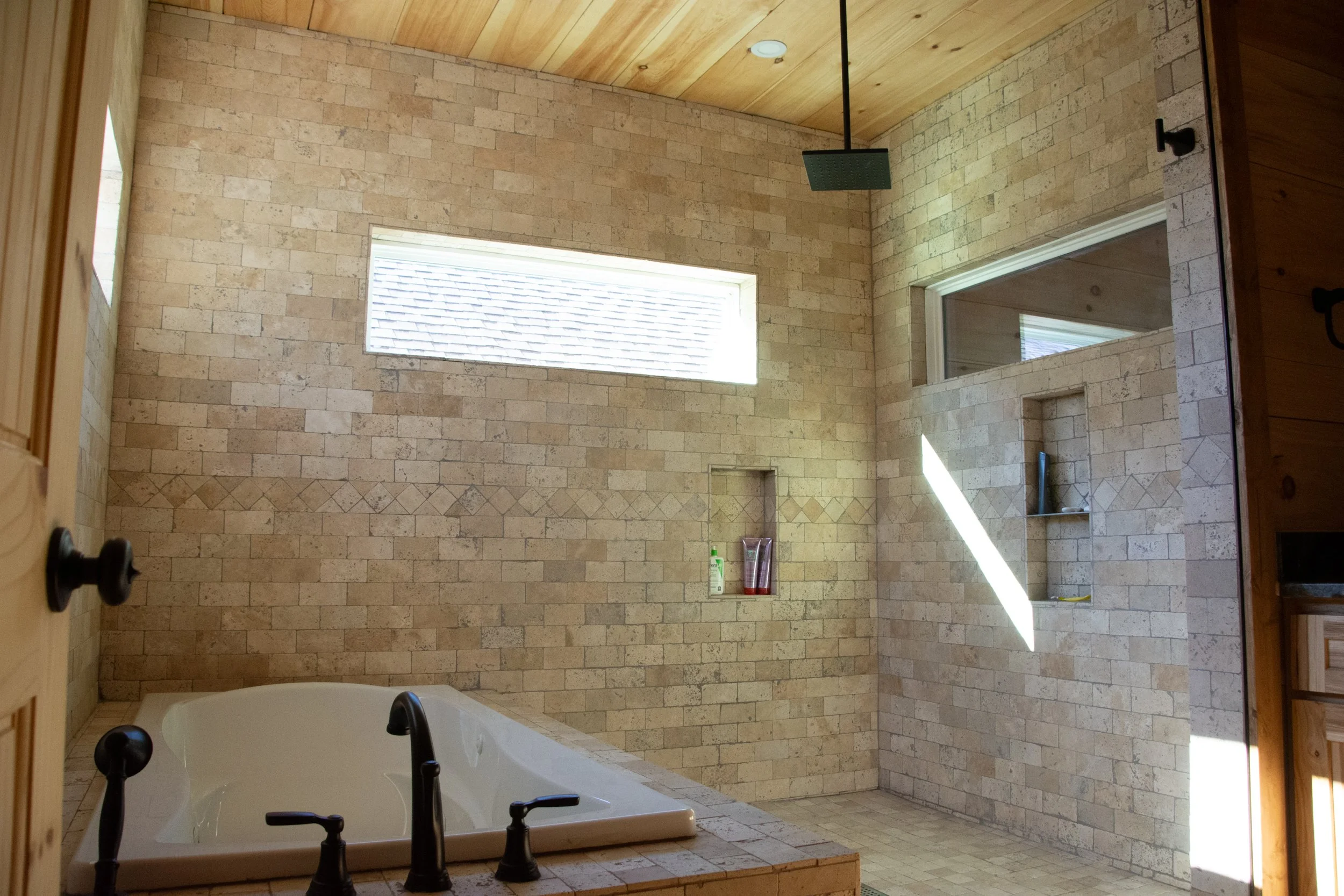 A spacious bathroom with a bathtub, brick-style tiled walls, a wooden ceiling, and a glass block window. There is a showerhead and built-in shelves holding toiletries. Natural light streams in, creating shadows on the walls.