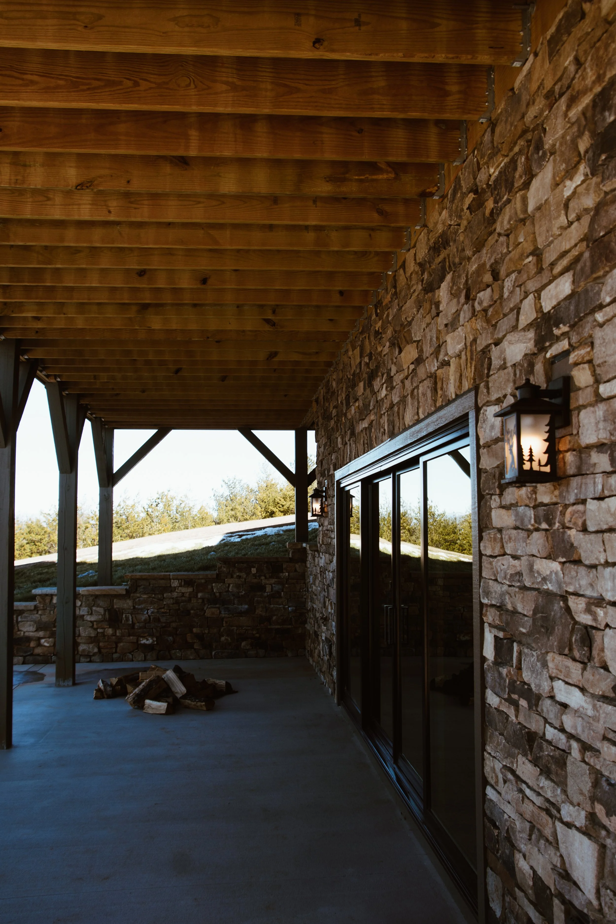 Covered outdoor patio area with wooden ceiling, stone wall, black sliding glass door, and wall lanterns, overlooking a grassy landscape.