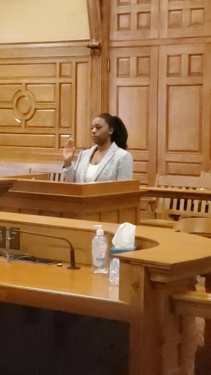 A stern woman with dark hair, wearing a light blazer, stands behind a wood podium speaking with her right arm raised.