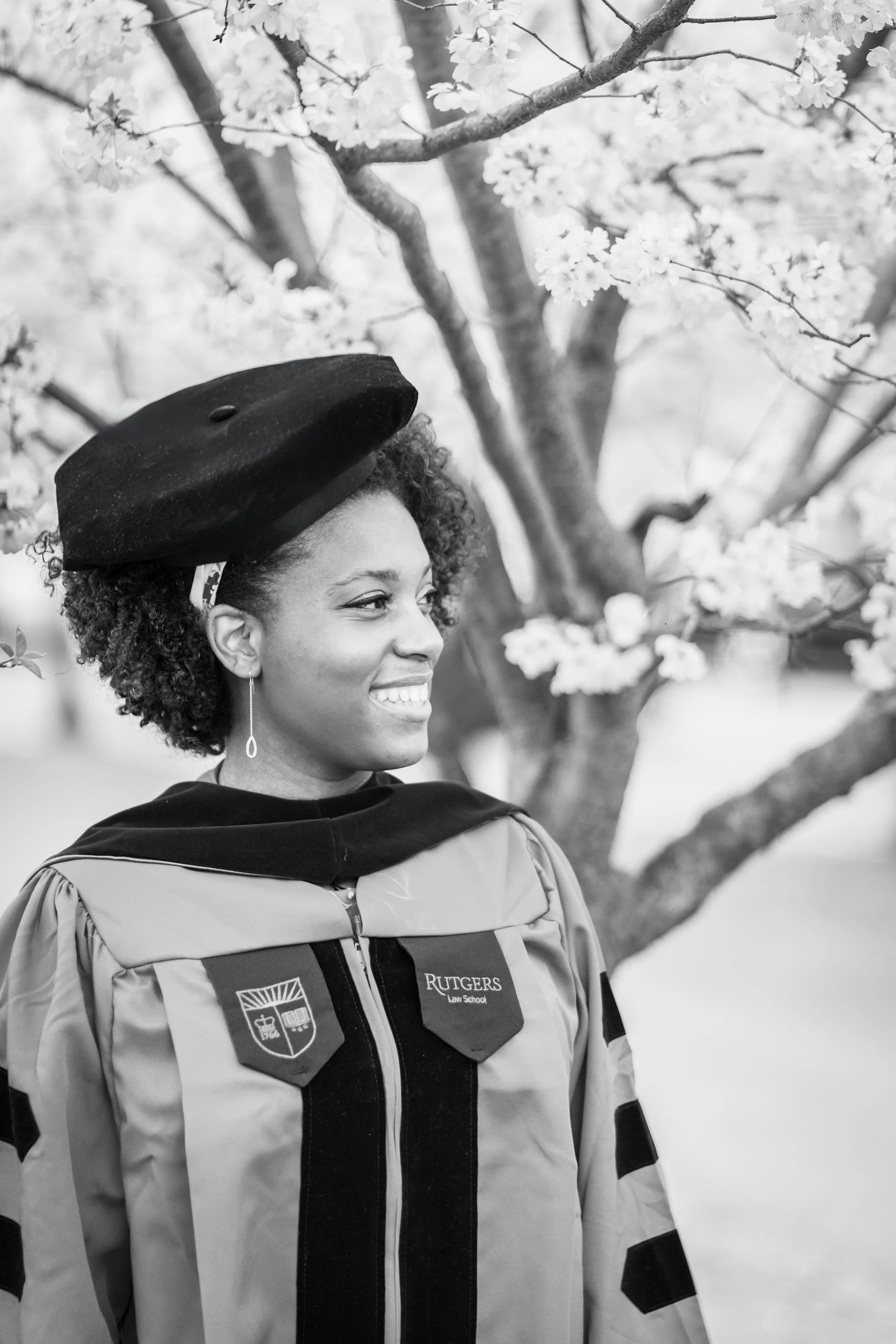 Black and white photo of a woman in law school graduation gown and cap, standing in front of a flowering tree, smiling.
