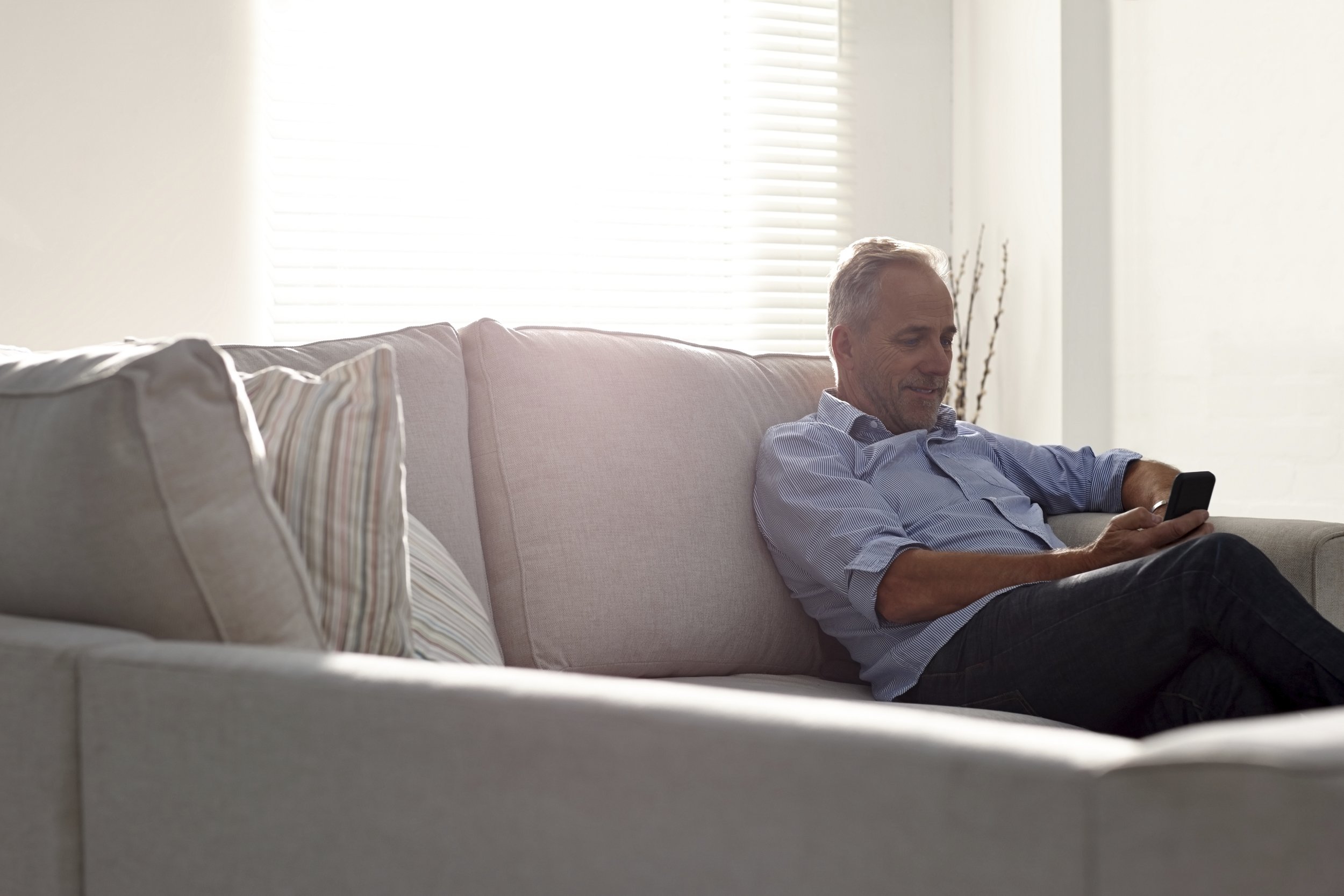 A middle-aged man sitting on a light-colored couch, looking at his smartphone in his hand, in a bright living room with sunlight coming through the window blinds.