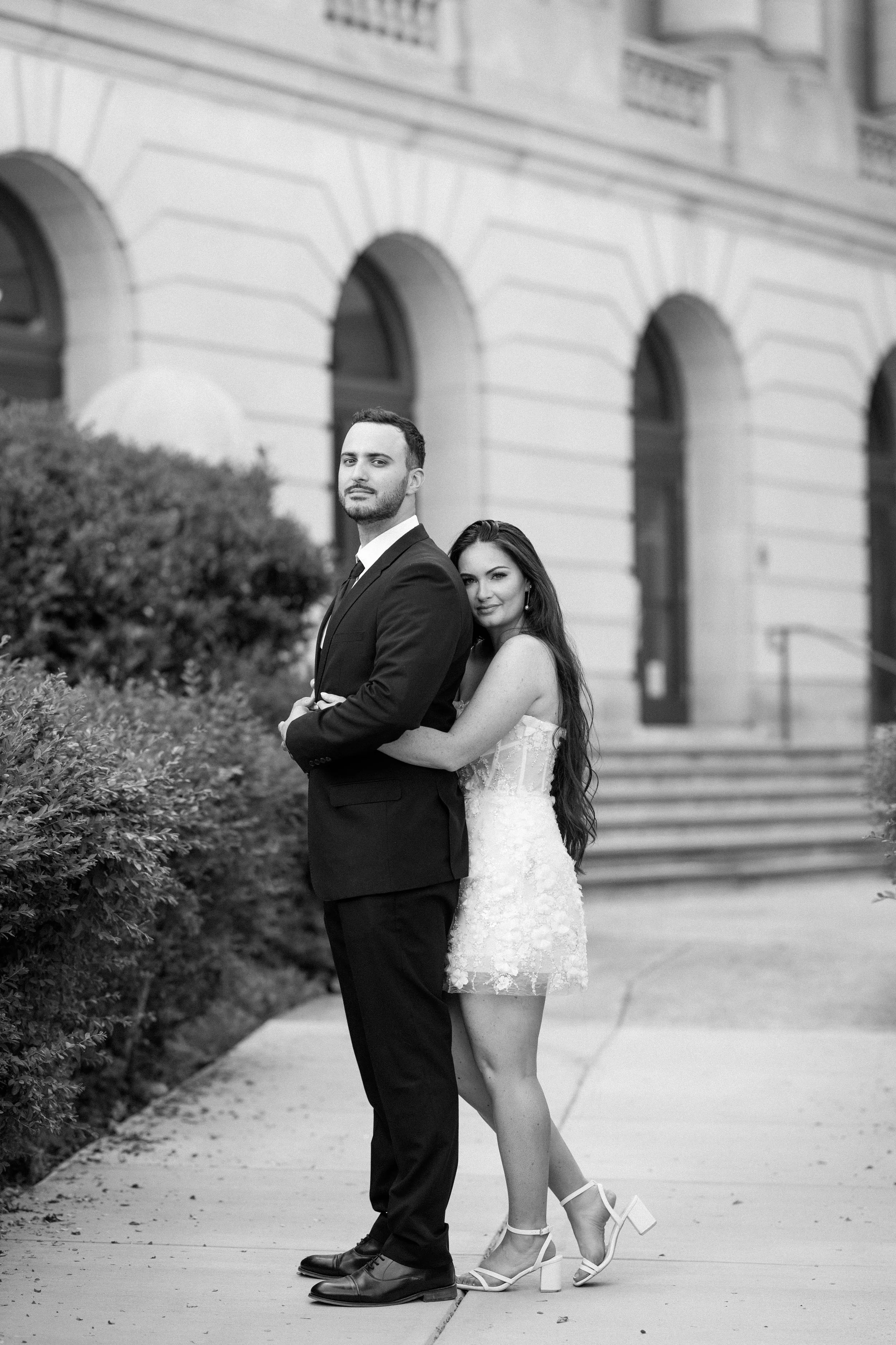 Black and white photo of a couple standing outdoors in front of a historic building with arched windows, with the woman hugging the man from behind on a sidewalk.