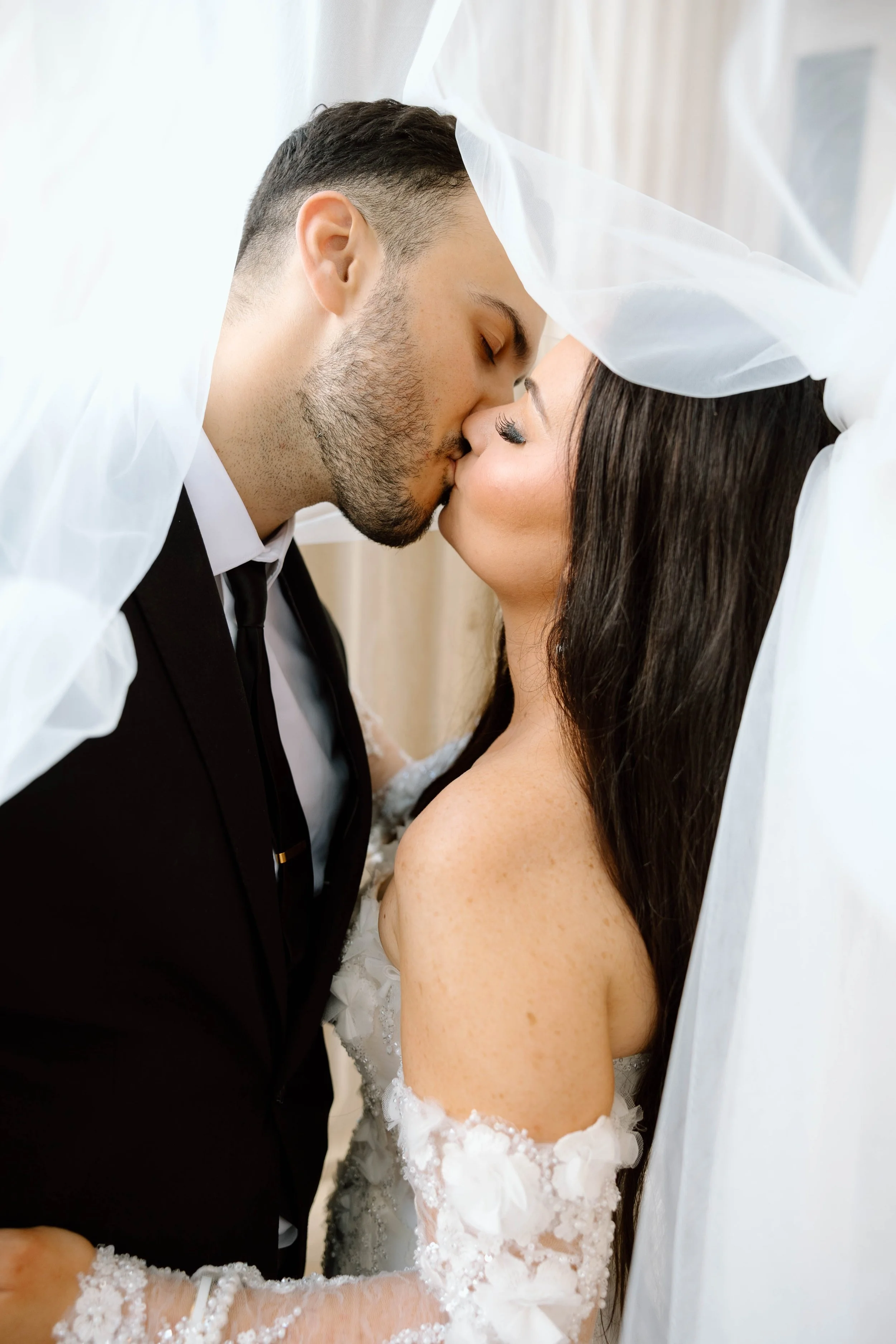 A couple in wedding attire sharing a kiss under a white veil.