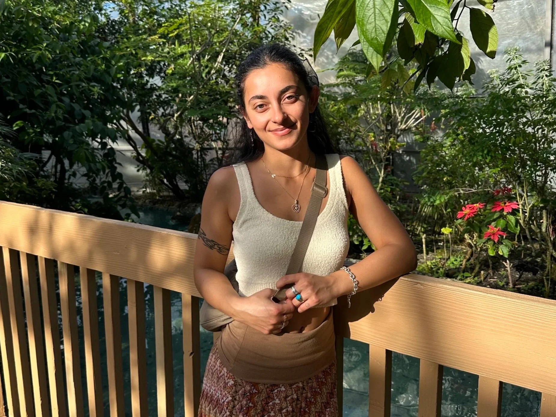 Young woman with dark hair wearing a beige tank top and patterned skirt, standing outdoors on a wooden bridge with lush green foliage and pink flowers in the background, smiling at the camera.