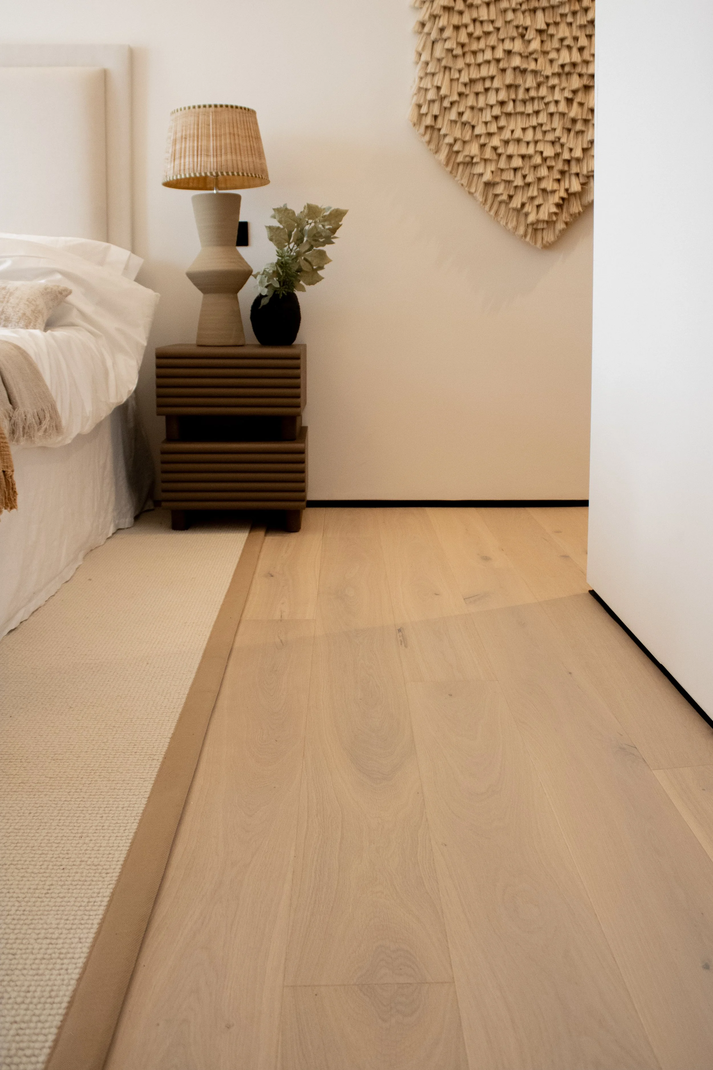 A bedroom corner with a bed, a nightstand, a lamp, a plant, a woven wall hanging, and light wooden flooring.