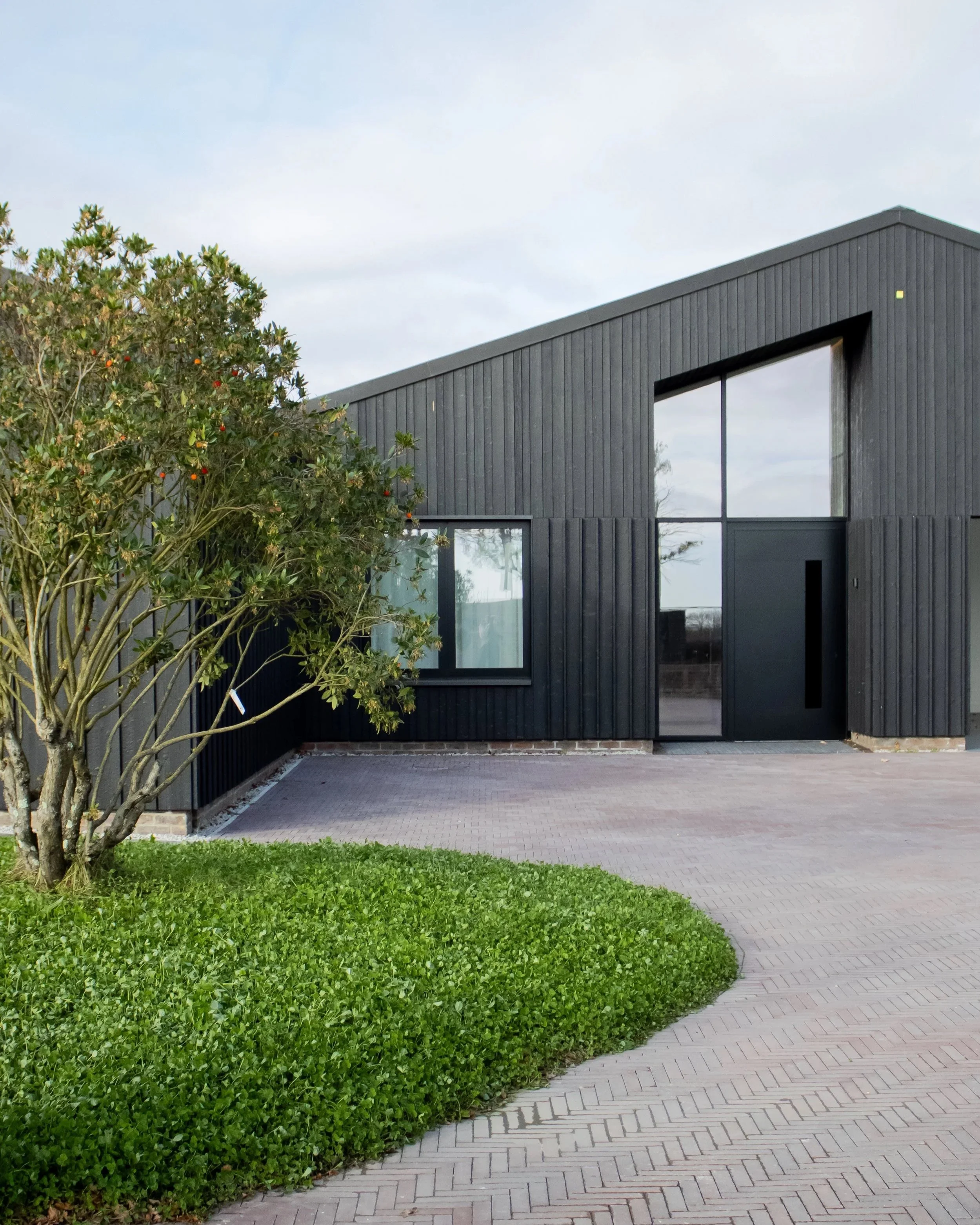 Modern black house with large glass windows and a curved brick walkway leading to the entrance, surrounded by a green lawn and a tree on the left.