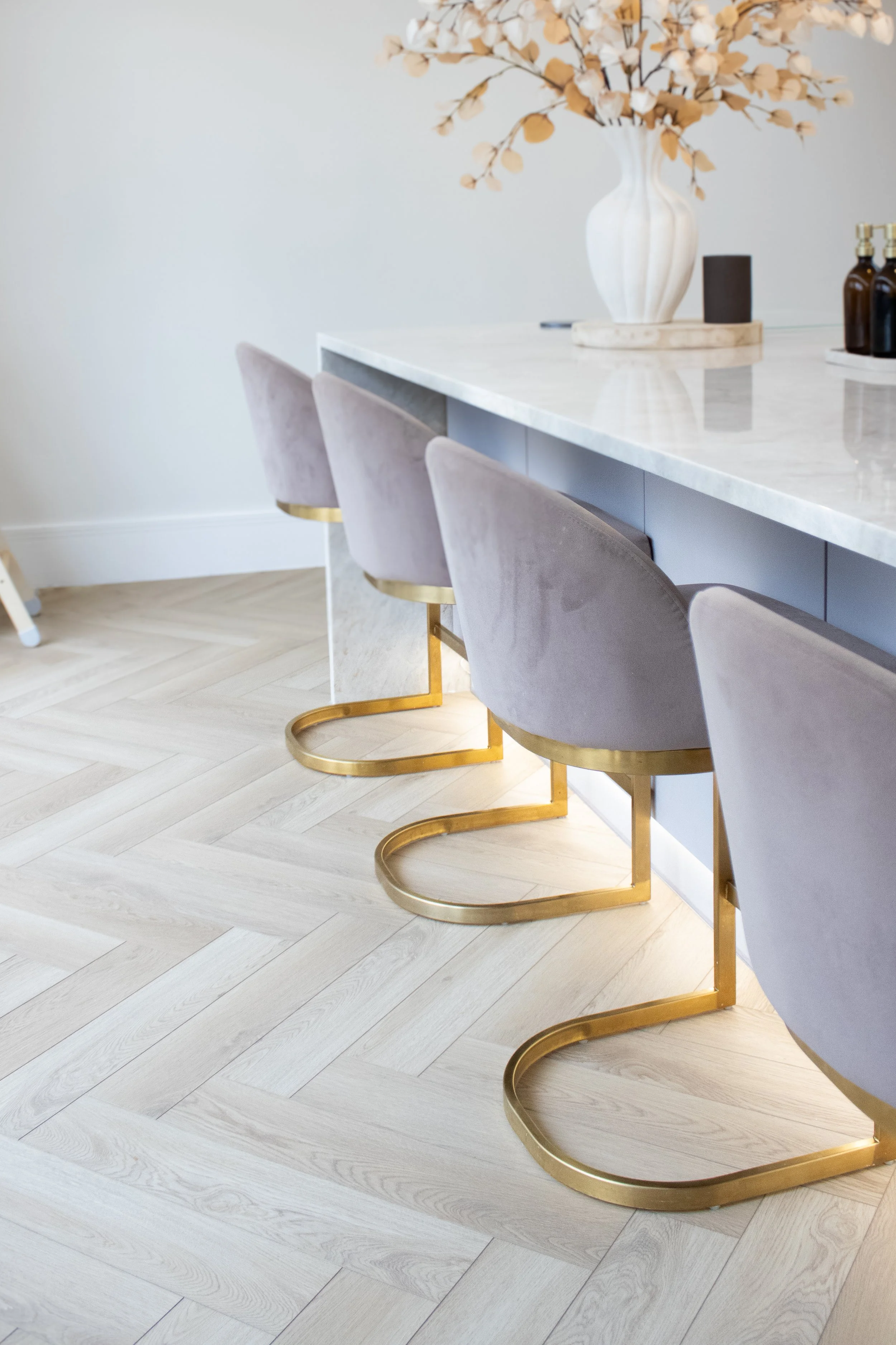 A row of four modern, plush, light grey bar stools with gold U-shaped bases at a white marble kitchen island with a minimalist decor, including a white vase with dried branches, amber bottles, and a black container.