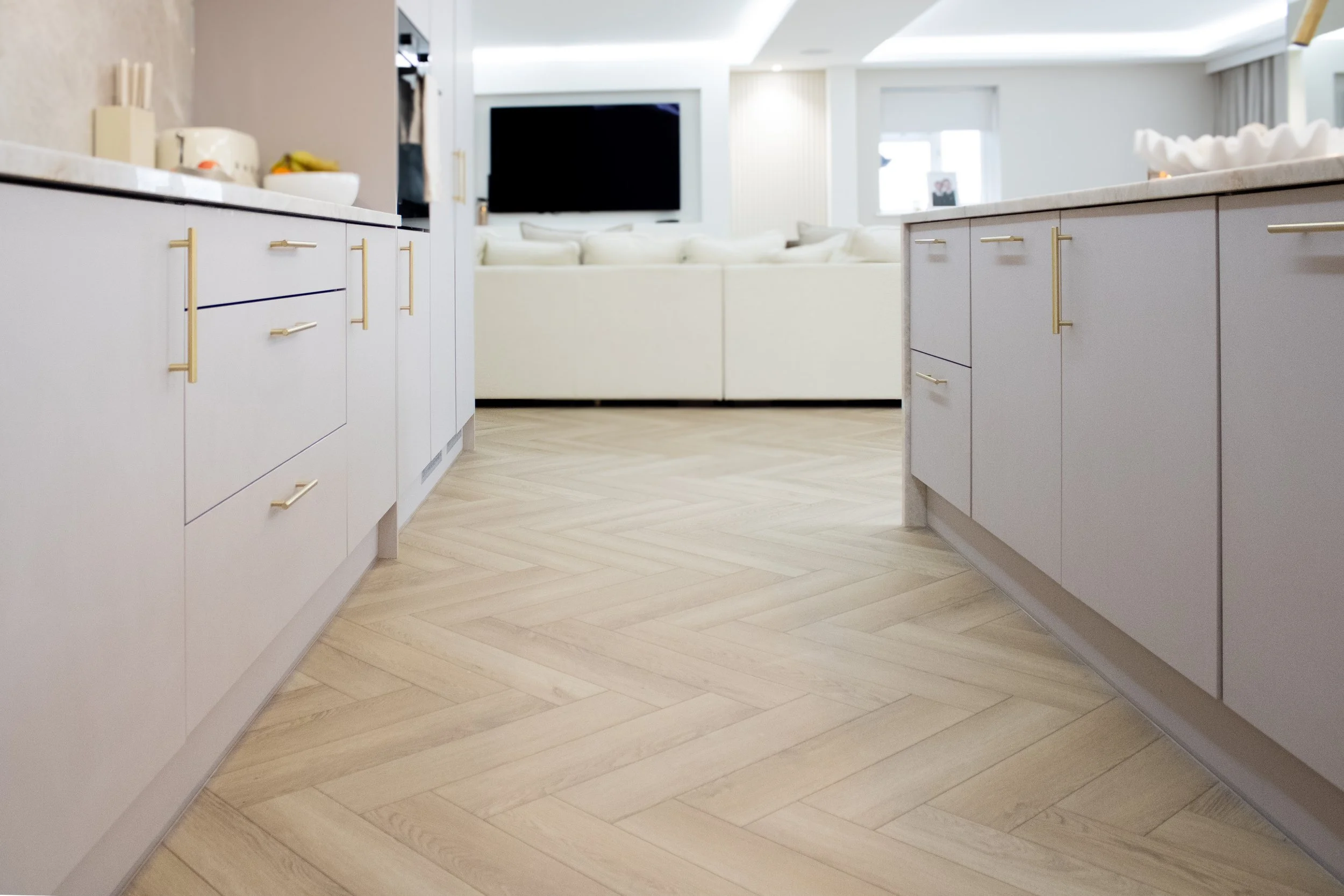 Kitchen with white cabinets and gold handles, light wood herringbone floor, view into a living room with white sofa and large television