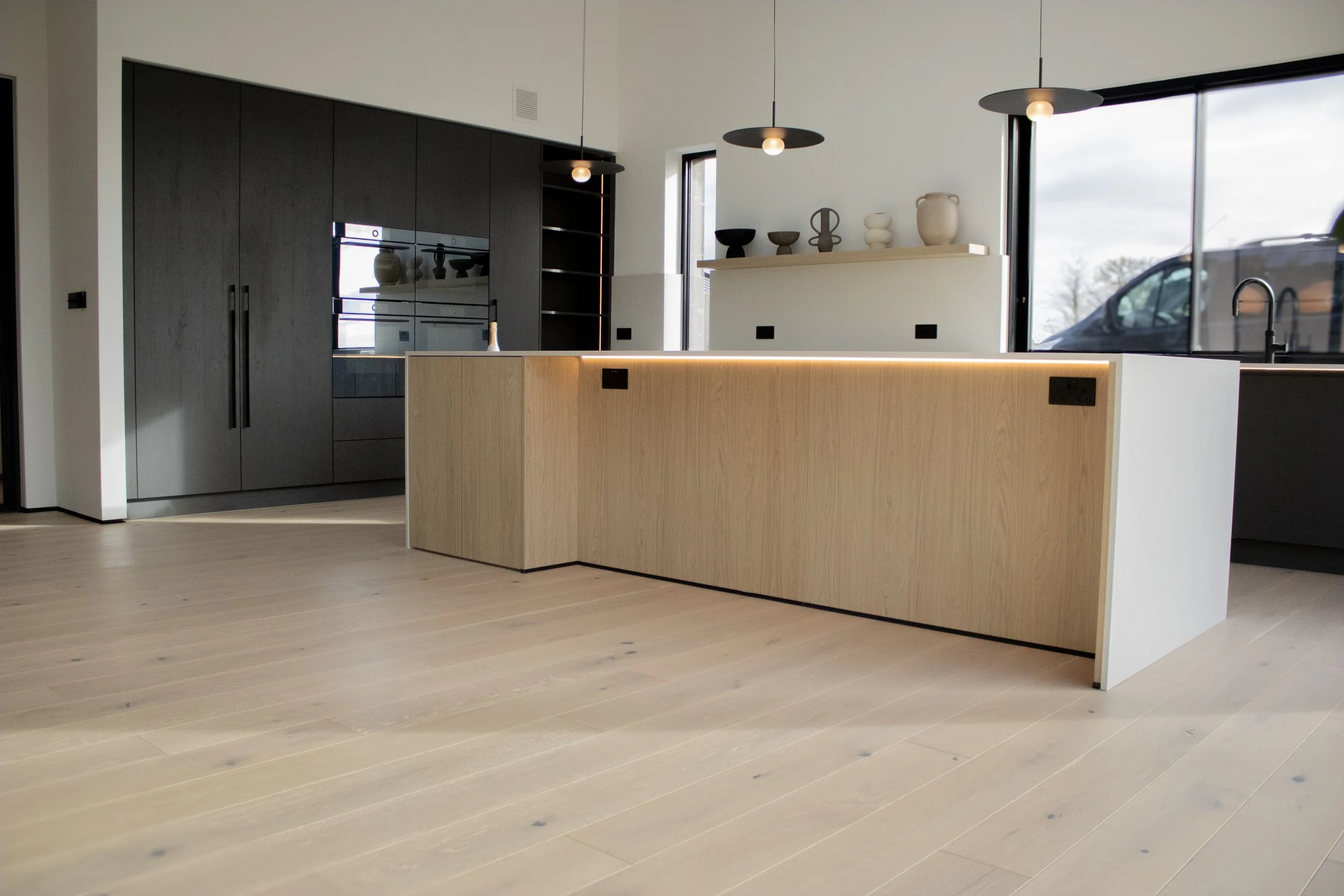Modern minimalist kitchen with light wood island, black cabinetry, and a large glass window letting in natural light.