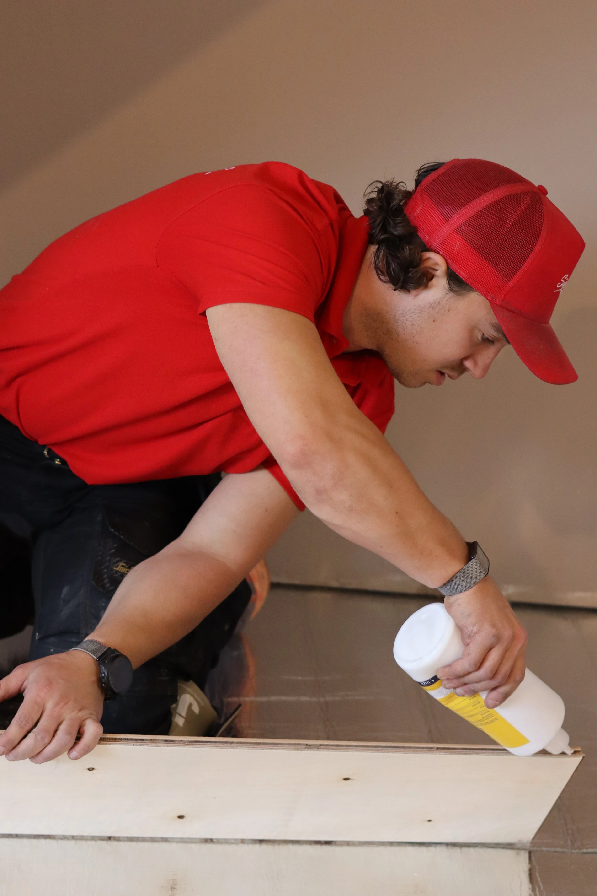 A man wearing a red cap and red shirt applying glue to a piece of wood for construction.