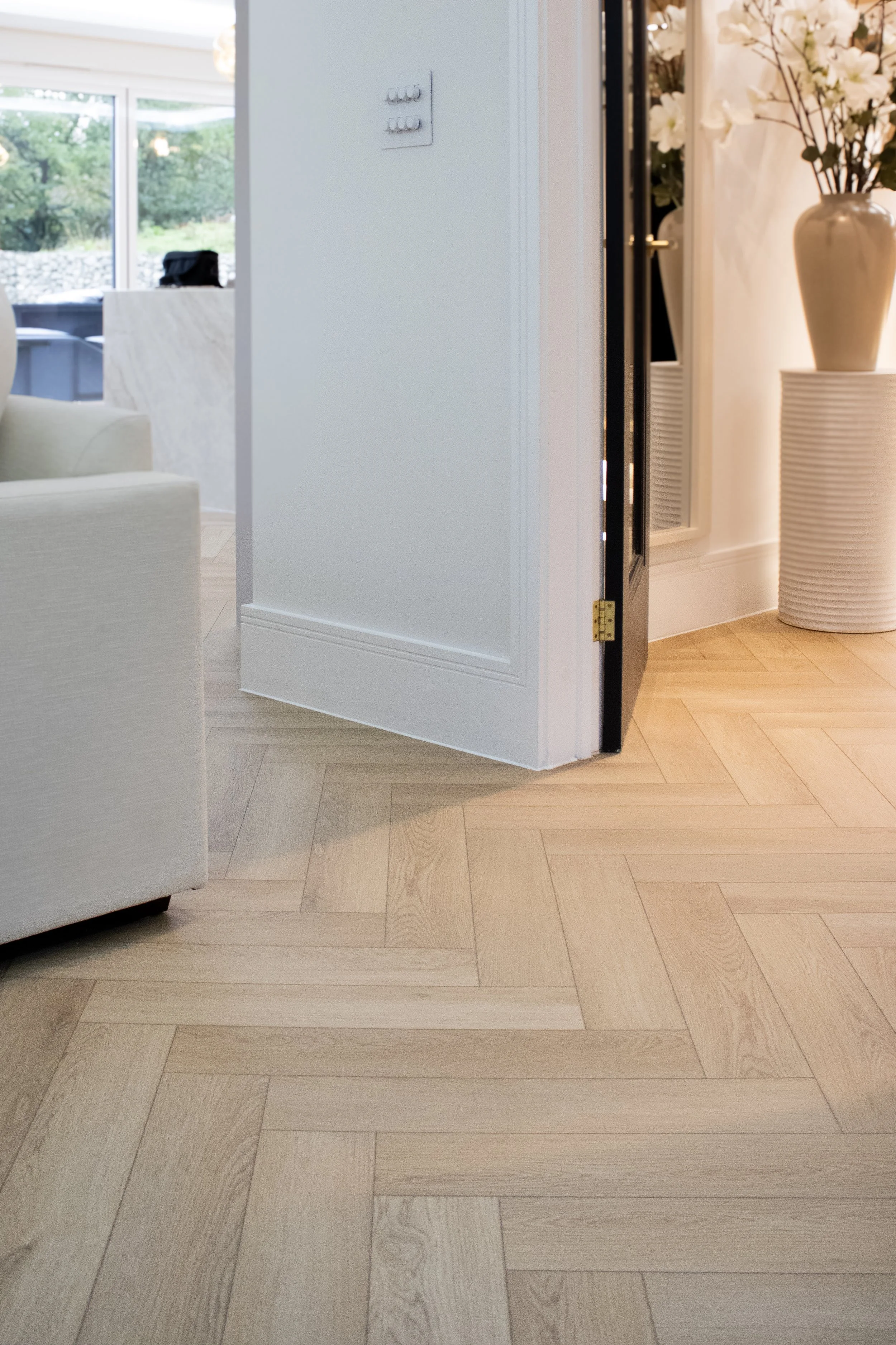 Interior view of a room with light wood herringbone patterned flooring, white walls, a beige sofa, and a large cream-colored vase with flowers.