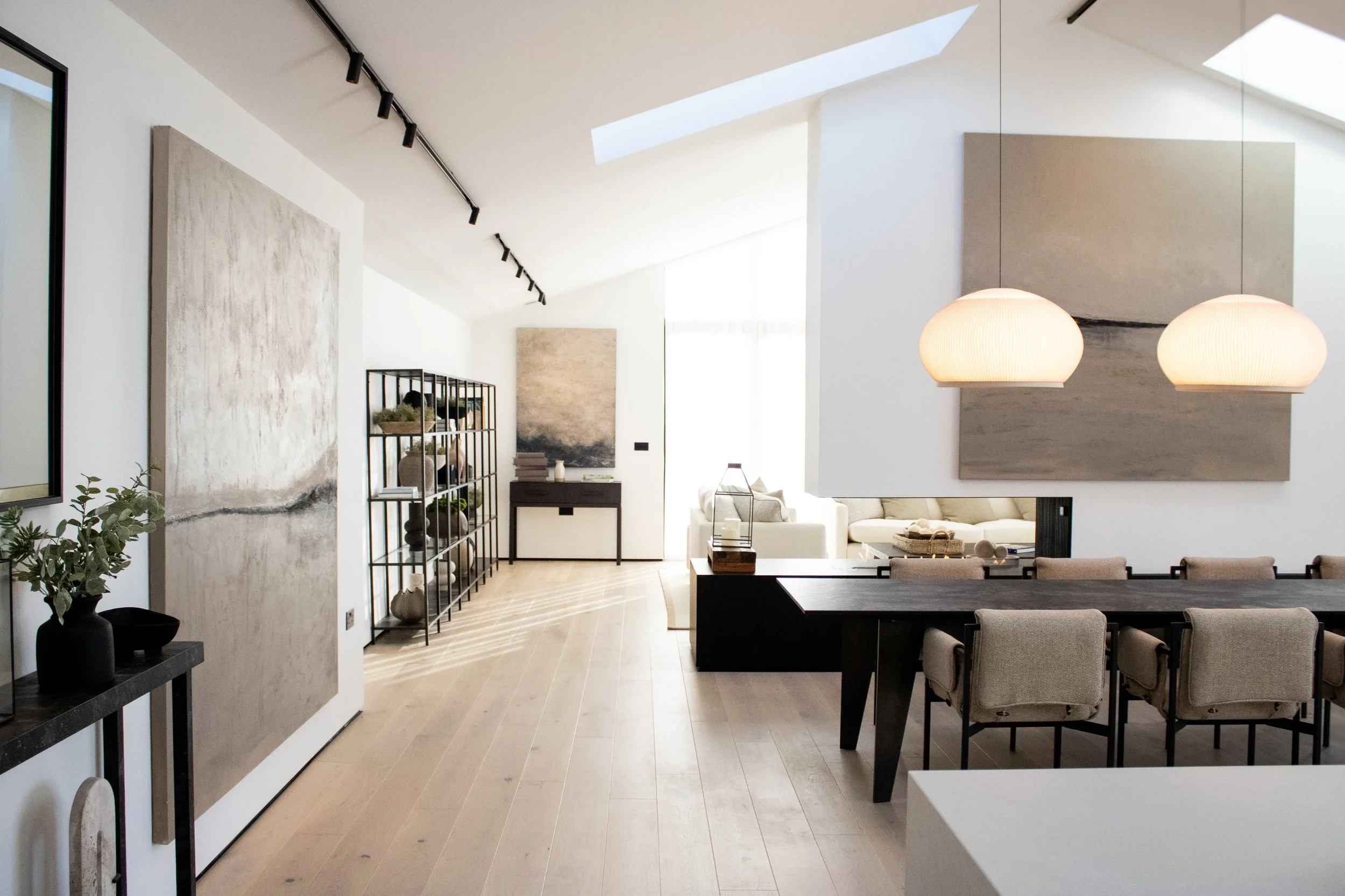 Modern open-concept living and dining area with light wood flooring, white walls, large abstract artwork, beige dining chairs, and a black dining table, illuminated by large hanging pendant lights.