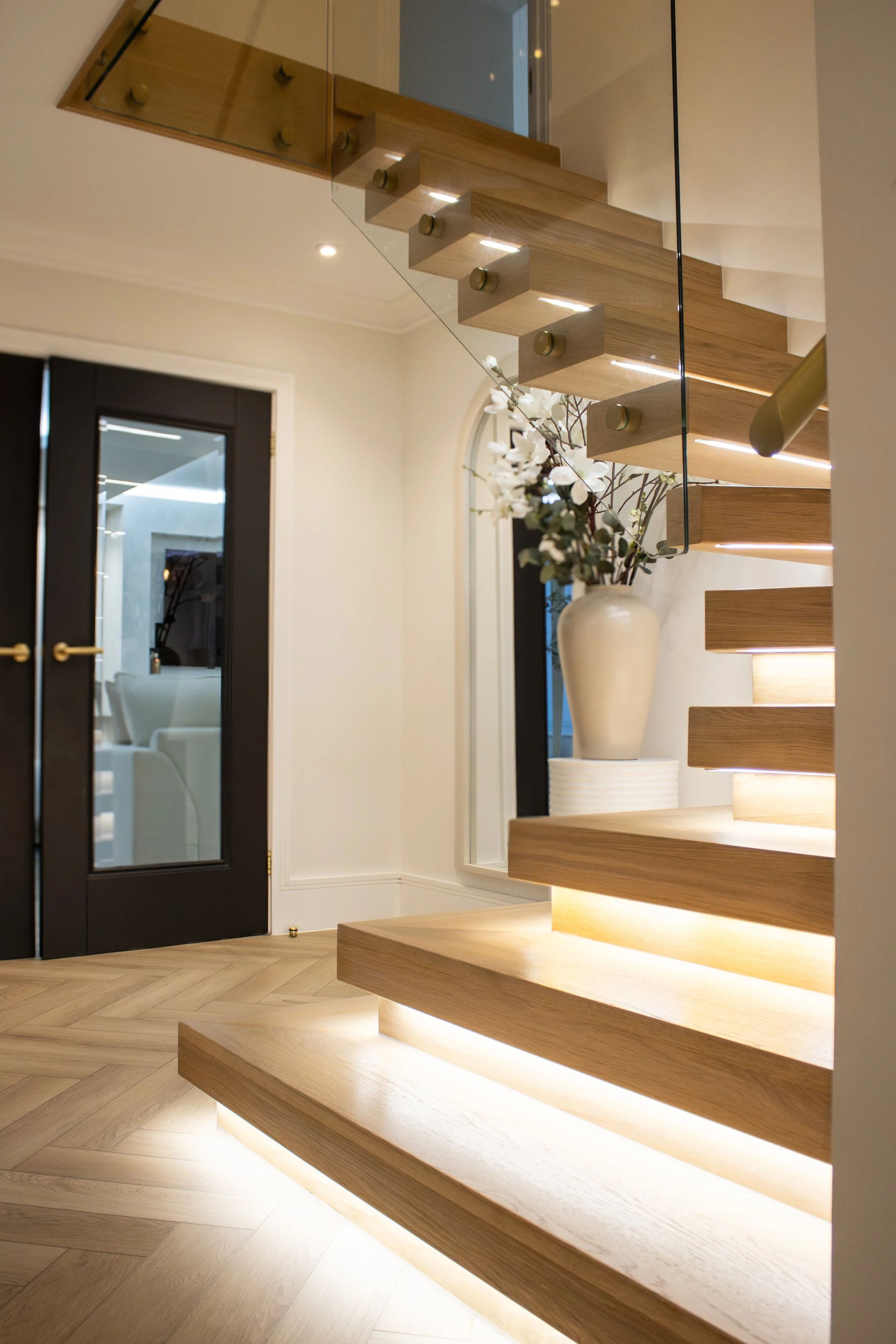 Modern interior with wooden staircase illuminated with built-in lights, a large white vase with white flowers, glass walls, black door with glass panel, and a mirror on the wall.