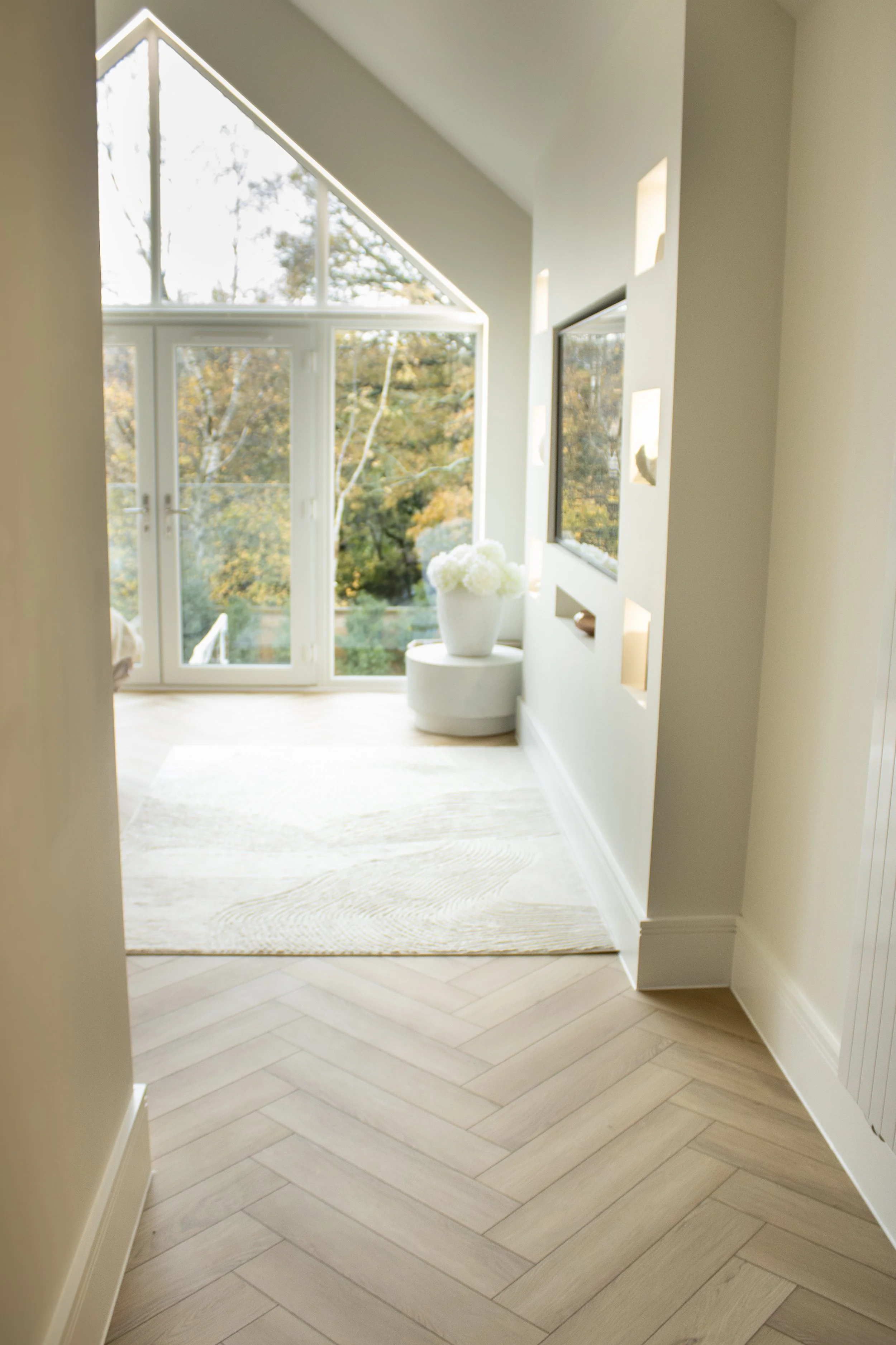 Bright, airy room with large windows and a glass door, featuring light-colored tiffany floors and a white rug, decorated with a large white vase of flowers and small wall niches.